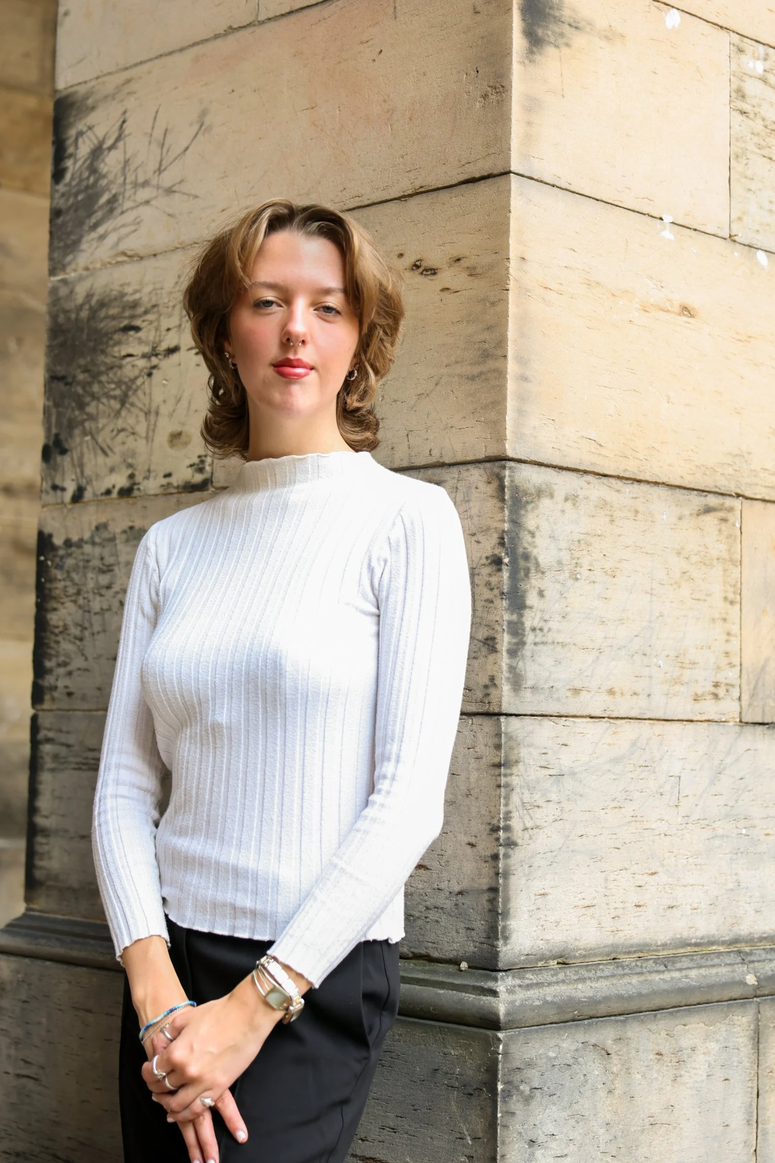 A young woman with short curly brown hair, wearing a white ribbed sweater and black pants, standing with crossed hands in front of a stone building.