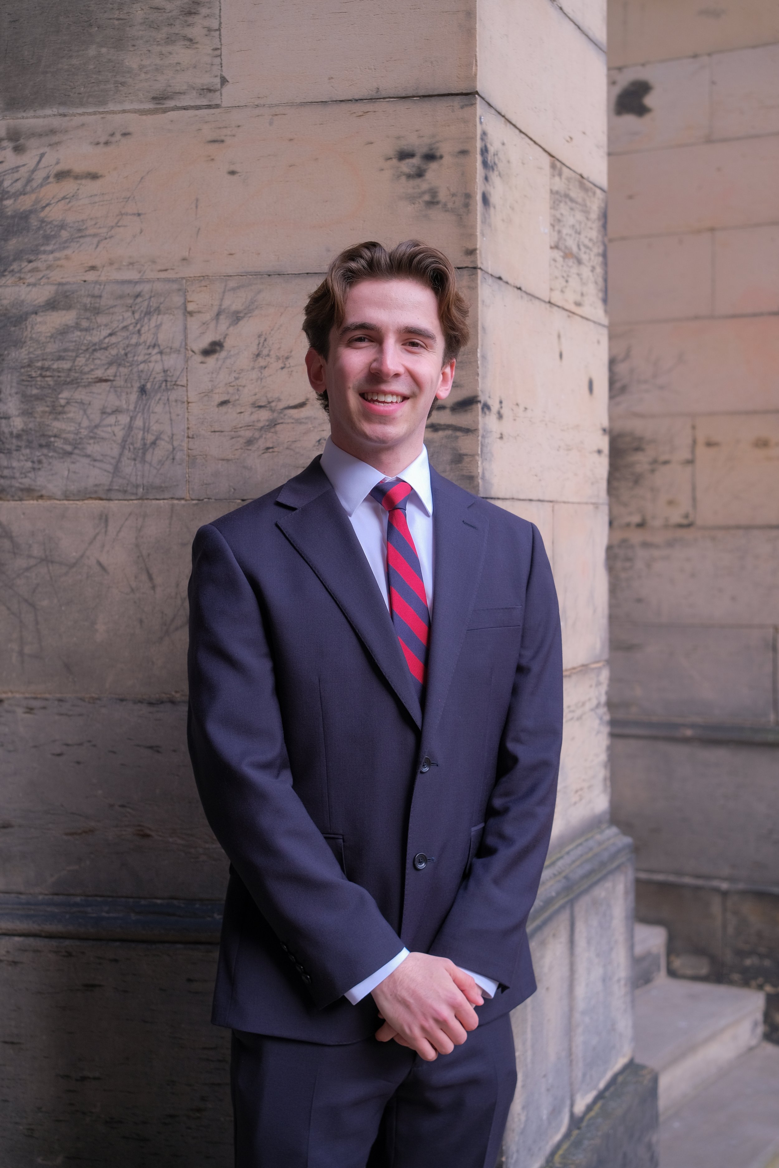 A young man in a navy suit with a white shirt and a red striped tie, standing against a stone wall with a smile.