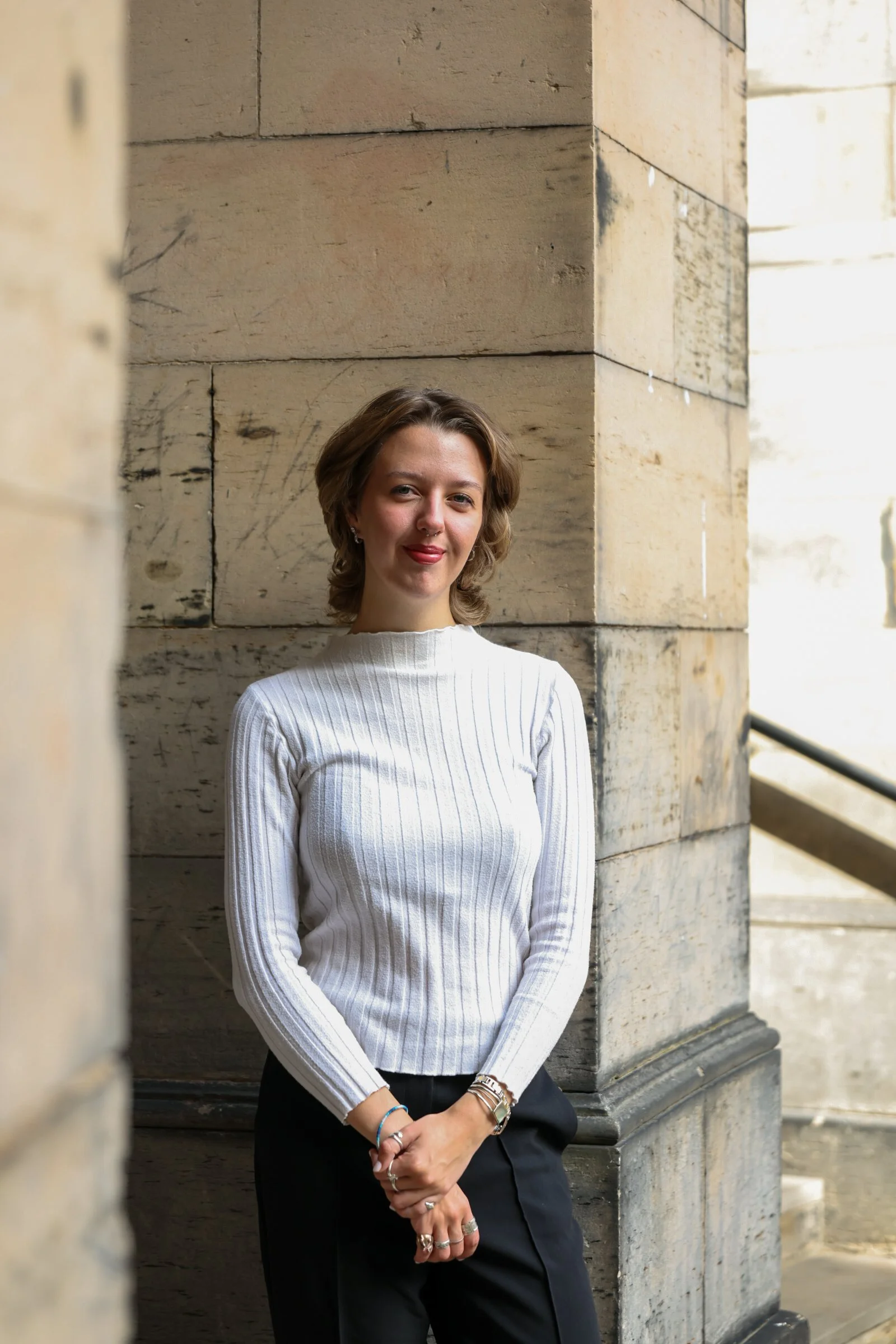 A young woman with short brown hair, wearing a white ribbed long-sleeve shirt and black pants, standing against a stone wall with an outdoor architectural setting.