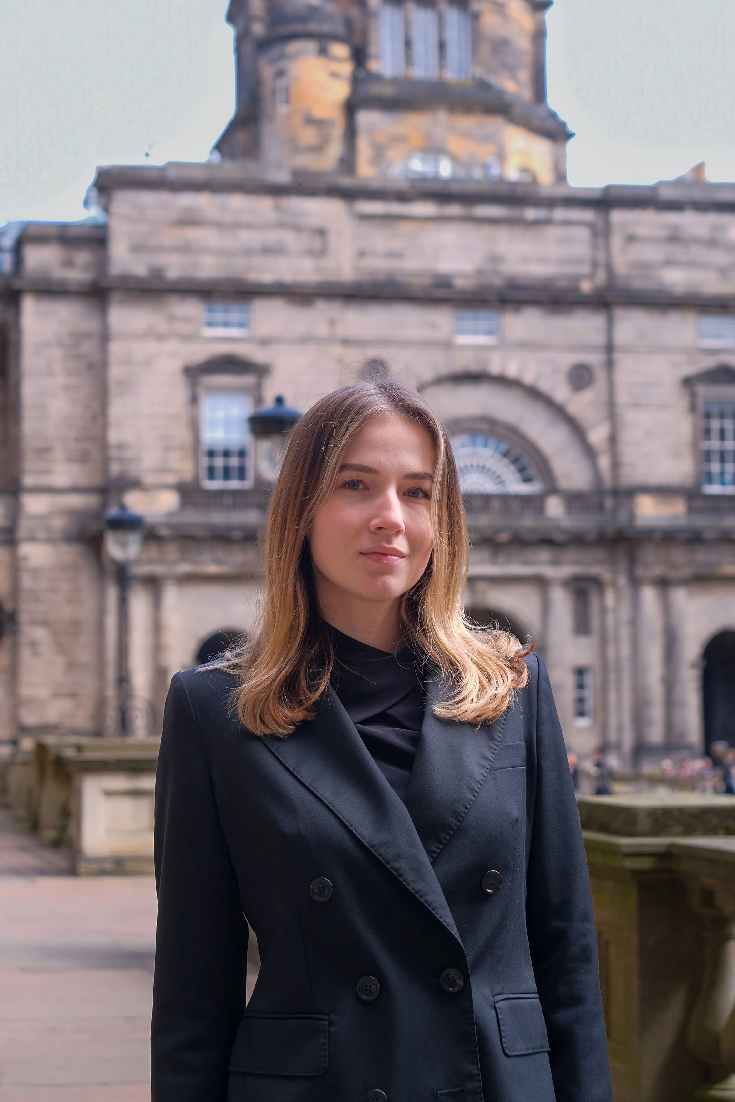 A young woman with shoulder-length brown hair standing in front of an old stone building with arched windows and a clock tower.