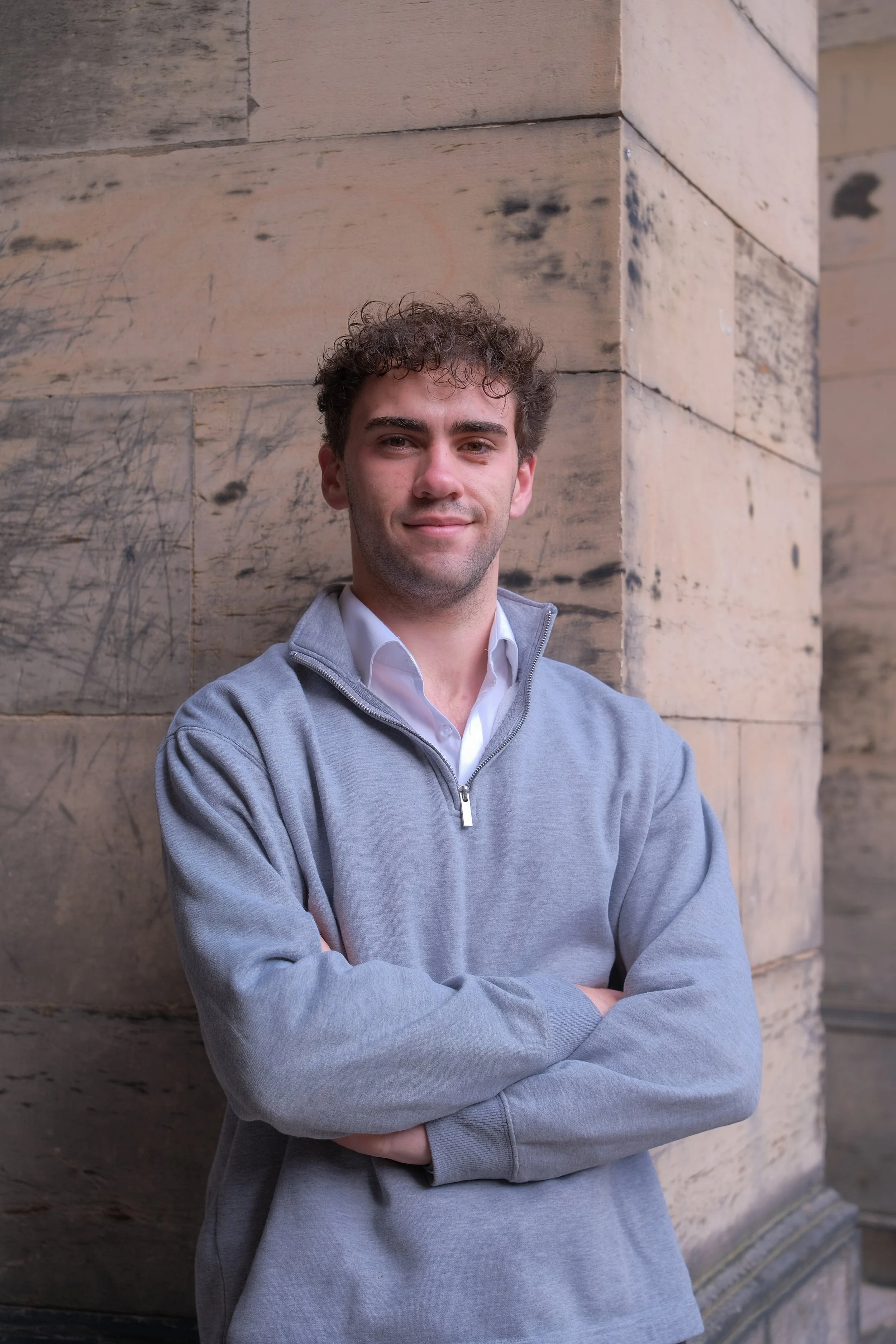 A young man with curly hair wearing a light gray zip-up sweater over a white collared shirt, standing outdoors against a beige stone wall with crossed arms and a slight smile.