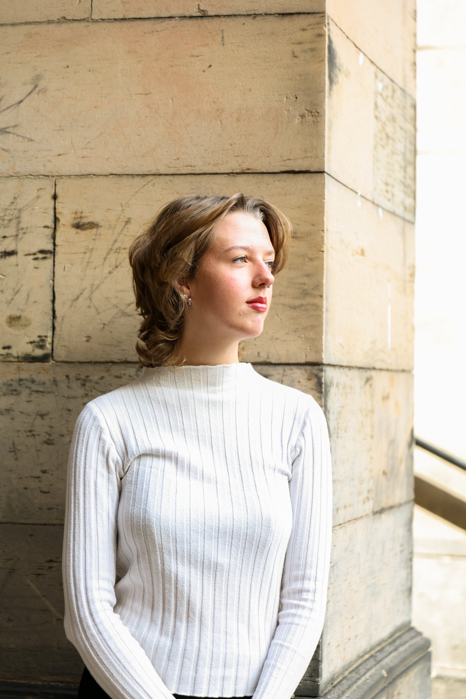 A young woman with shoulder-length light brown hair, dressed in a white ribbed turtleneck sweater, standing outdoors against a stone wall, gazing thoughtfully into the distance.
