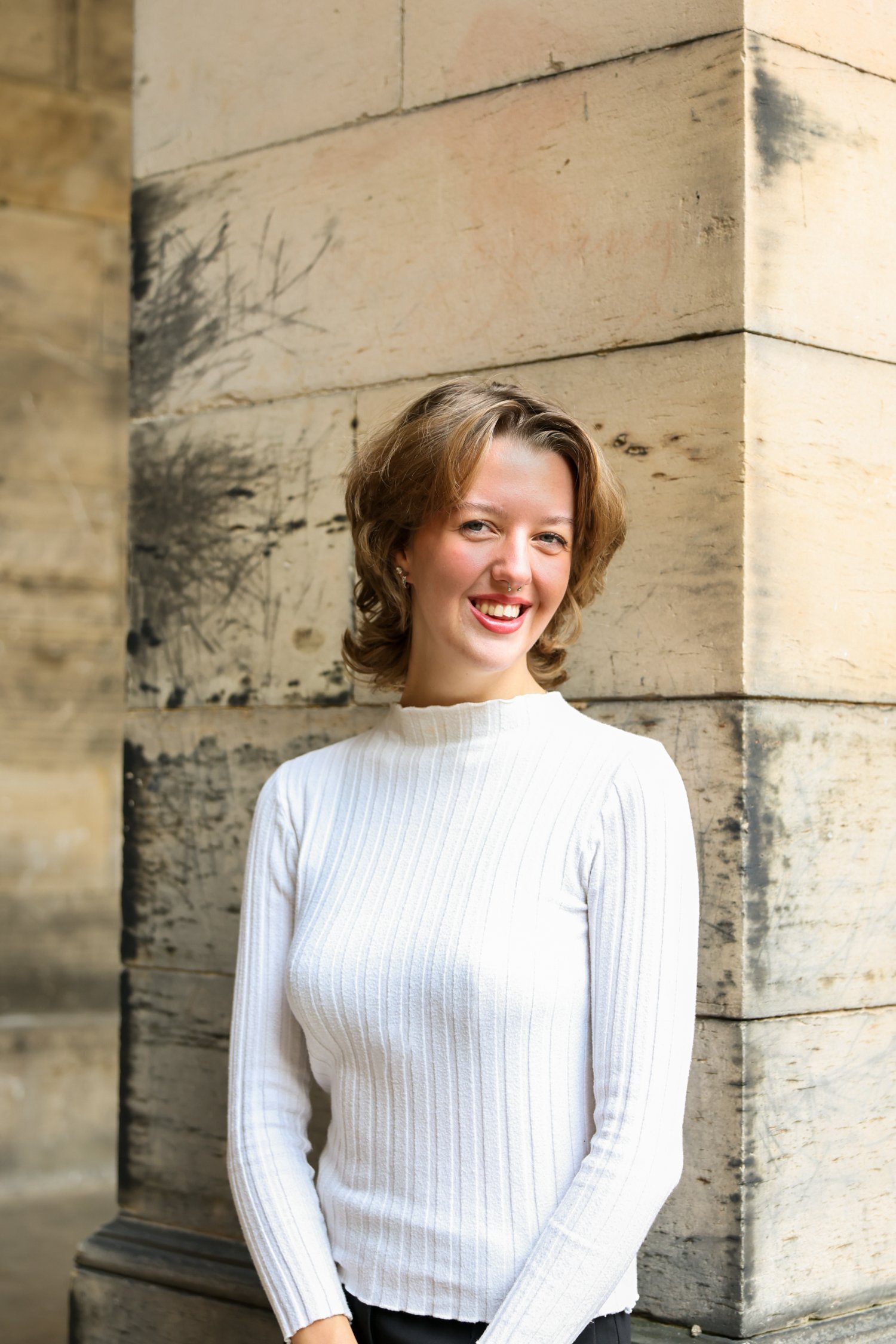 Smiling woman with short, wavy brown hair, wearing a white ribbed sweater, standing next to a stone building with weathered, light-colored bricks.