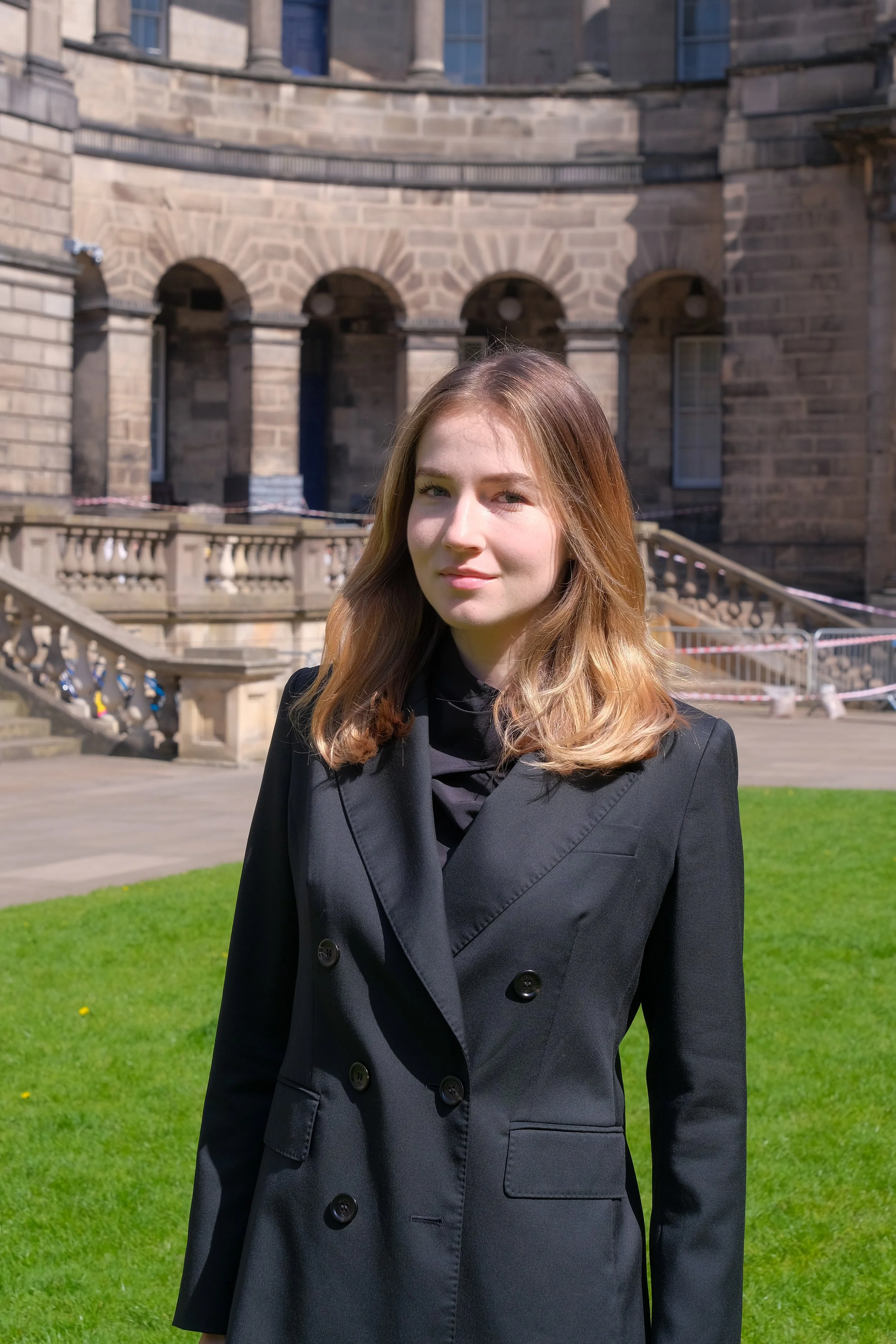 A young woman with shoulder-length blonde hair wearing a black double-breasted coat standing outside in front of a historic stone building with arches and a staircase.