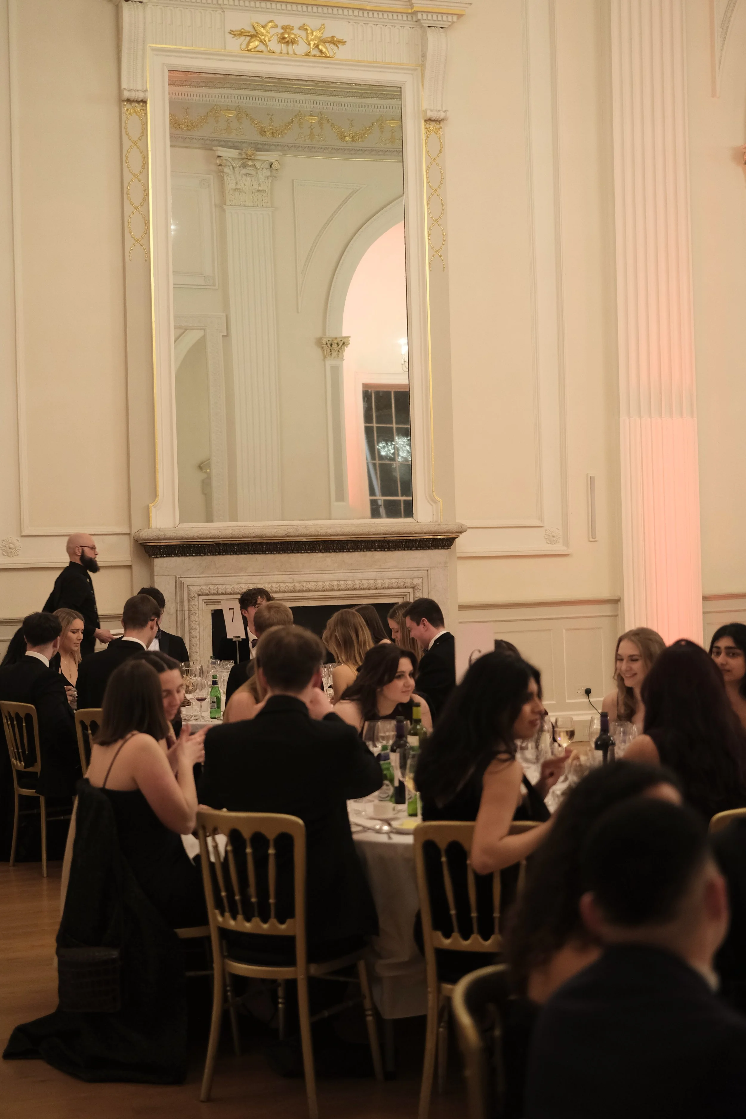 People dining at a formal event in a grand hall with elegant decor, a large mirror above a fireplace, and soft lighting.