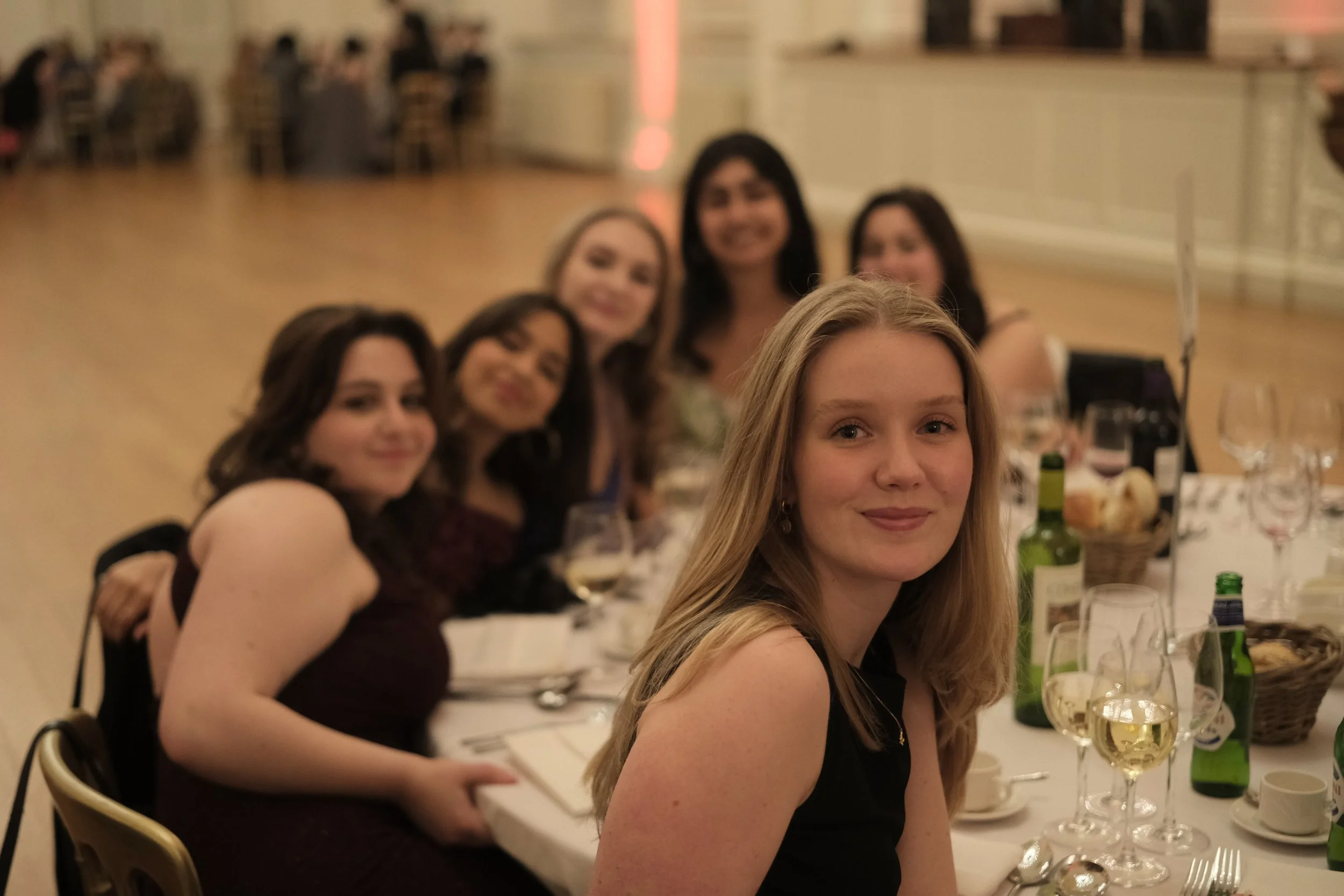 A group of women sitting at a long party table smiling at the camera, with glasses of wine and bottles on the table, in a decorated ballroom.