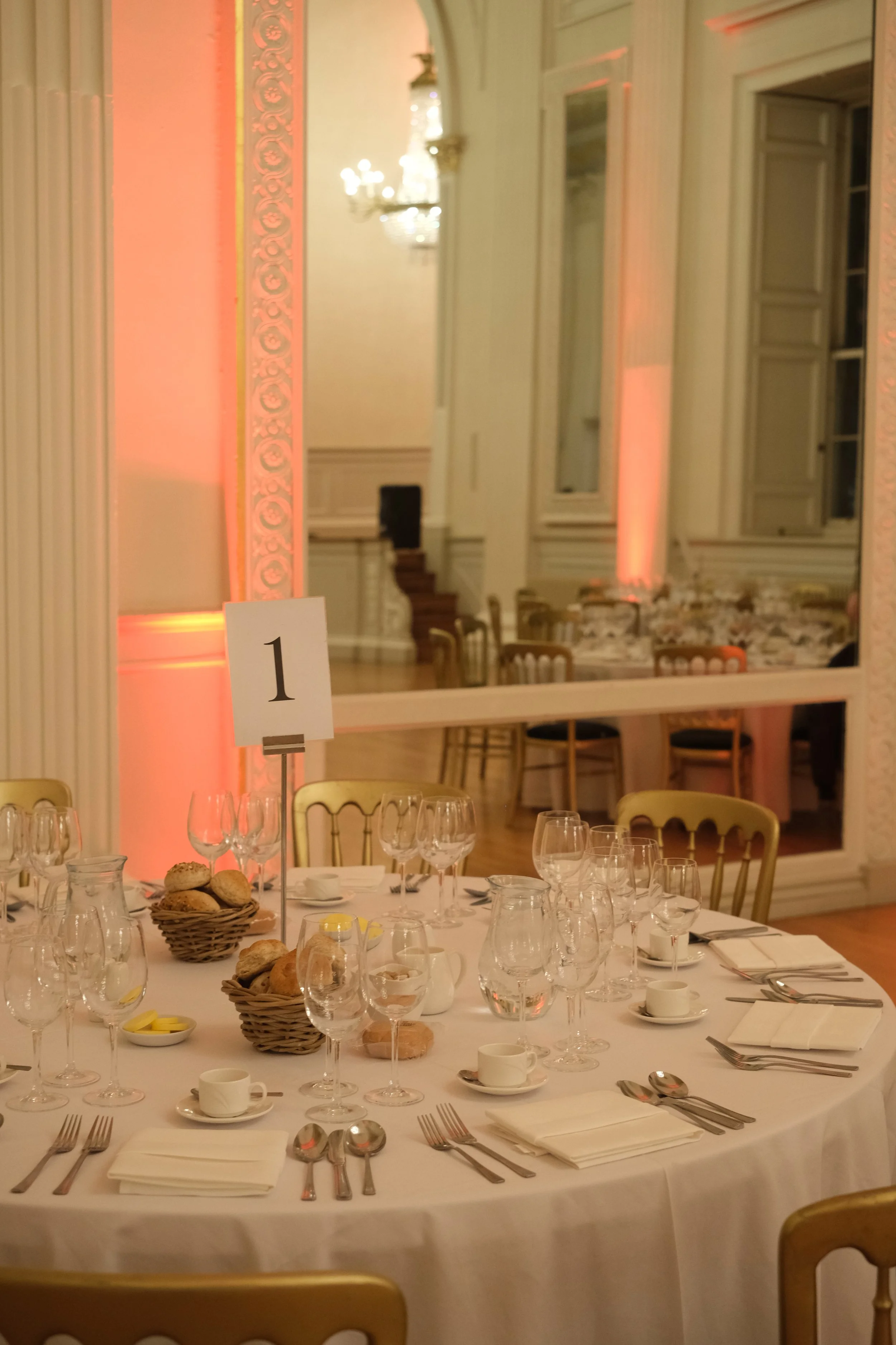 Elegant banquet table set with glassware, plates, silverware, cups, and napkins in a decorated room with warm lighting and a large mirror.