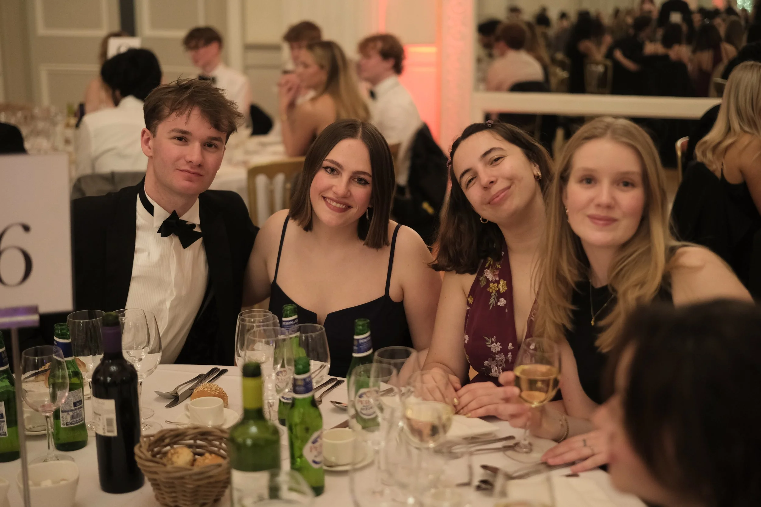 Group of four young people sitting at a dinner table during an event or celebration, smiling at the camera, with bottles of drinks and table settings visible.