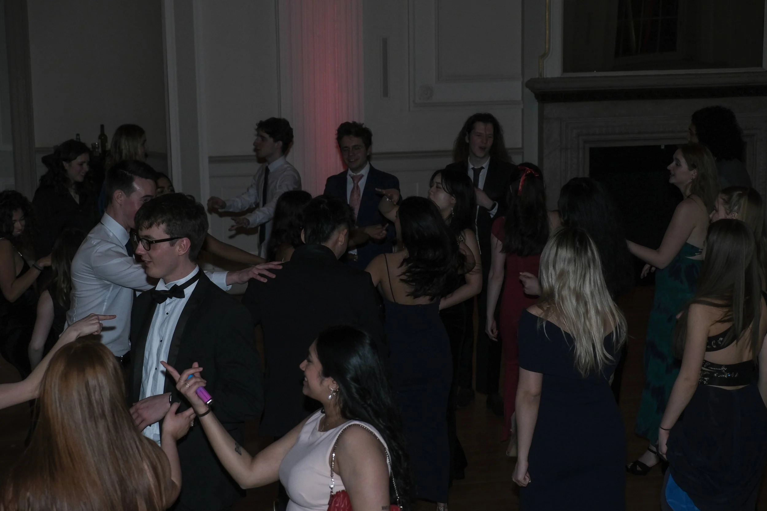 A group of young people dancing at a party or event in a dimly lit room with elegant decor and a fireplace.
