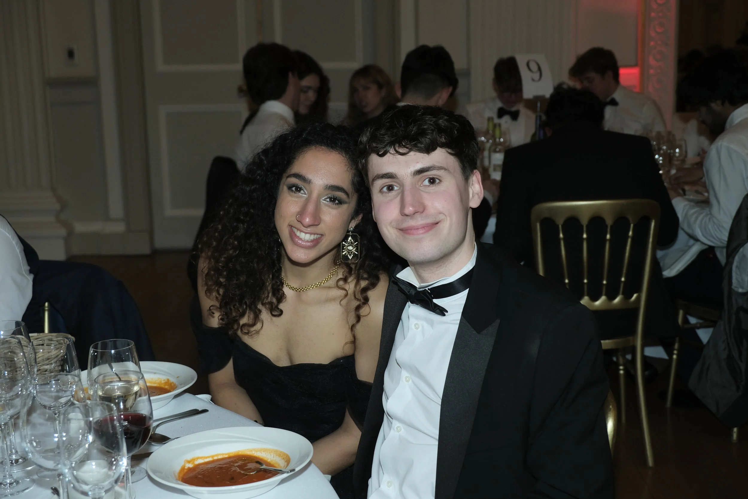 A young woman with dark curly hair and a dark dress, and a young man with short brown hair in a tuxedo, are sitting close together and smiling at the camera at a formal dinner event.