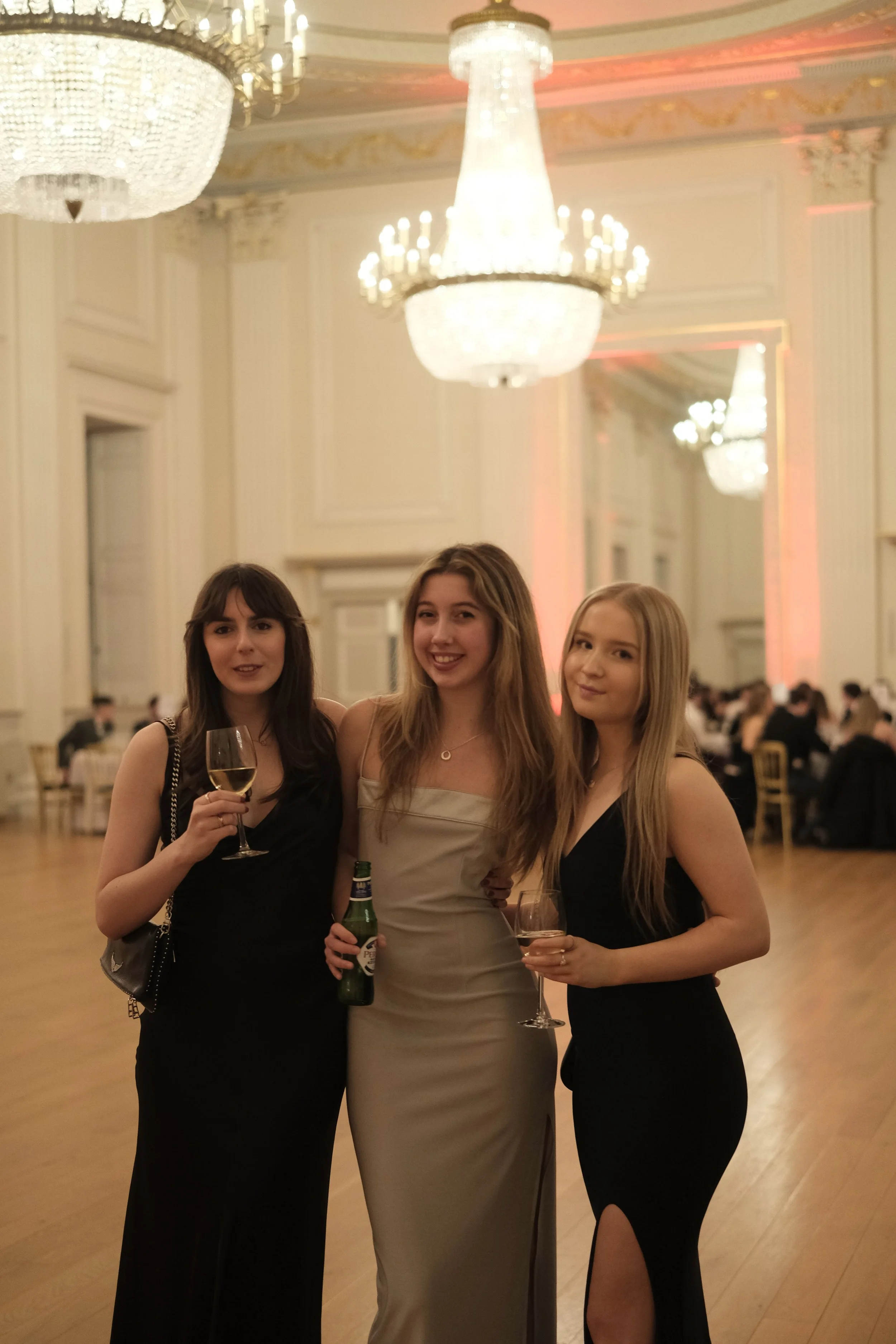 Three young women in elegant evening dresses holding drinks at a formal event in a grand ballroom with chandeliers and ornate decor.
