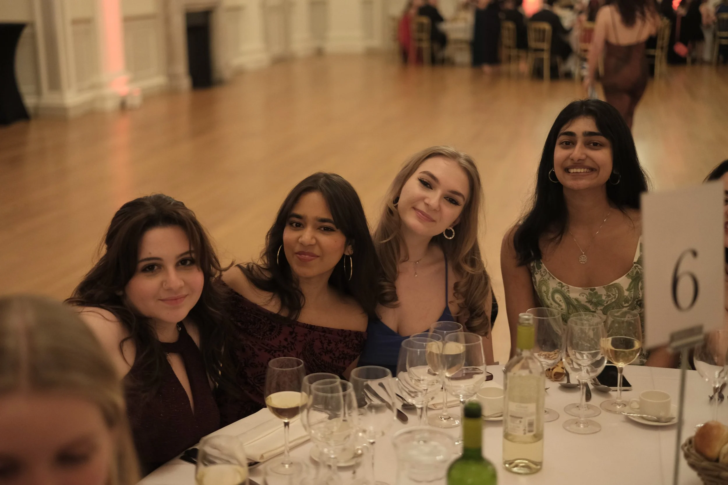 Four young women sitting at a formal dinner table with wine glasses, a bottle of wine, and bread rolls, smiling at the camera in an elegant event hall.