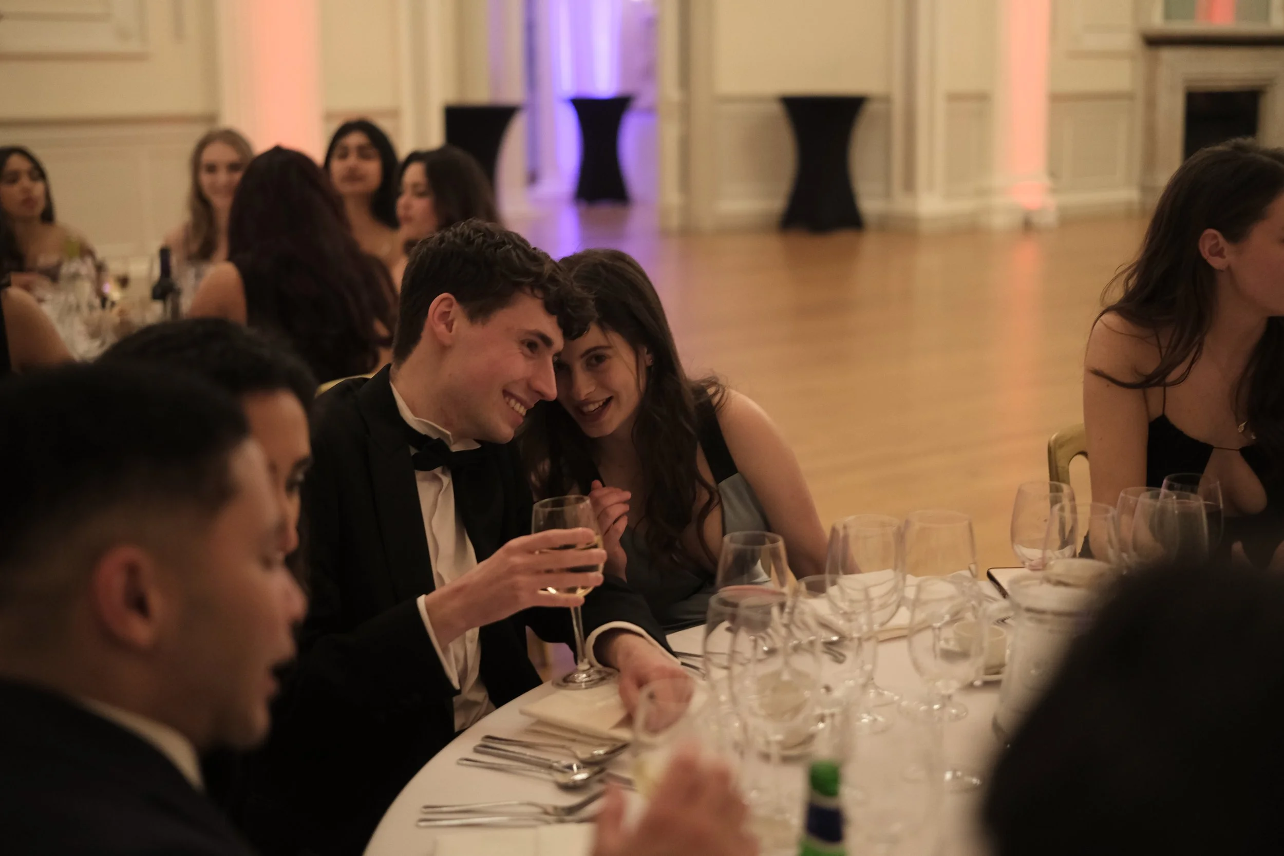 People dressed in formal attire sitting around a table at an elegant event, with two women whispering to each other, smiling.