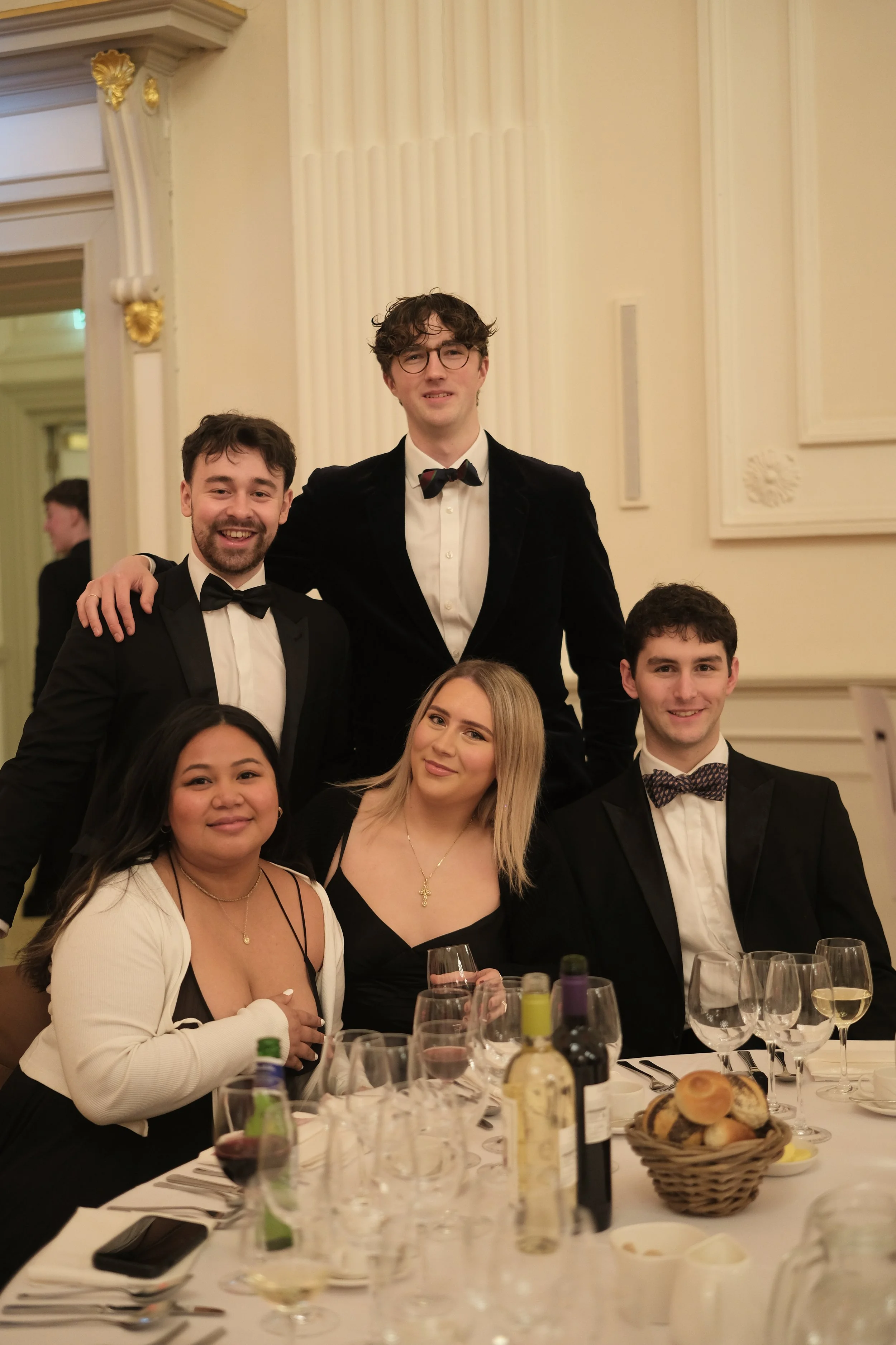 Group of six young adults in formal attire at a dinner party, posing for a photograph in an elegant room with ornate decorations.
