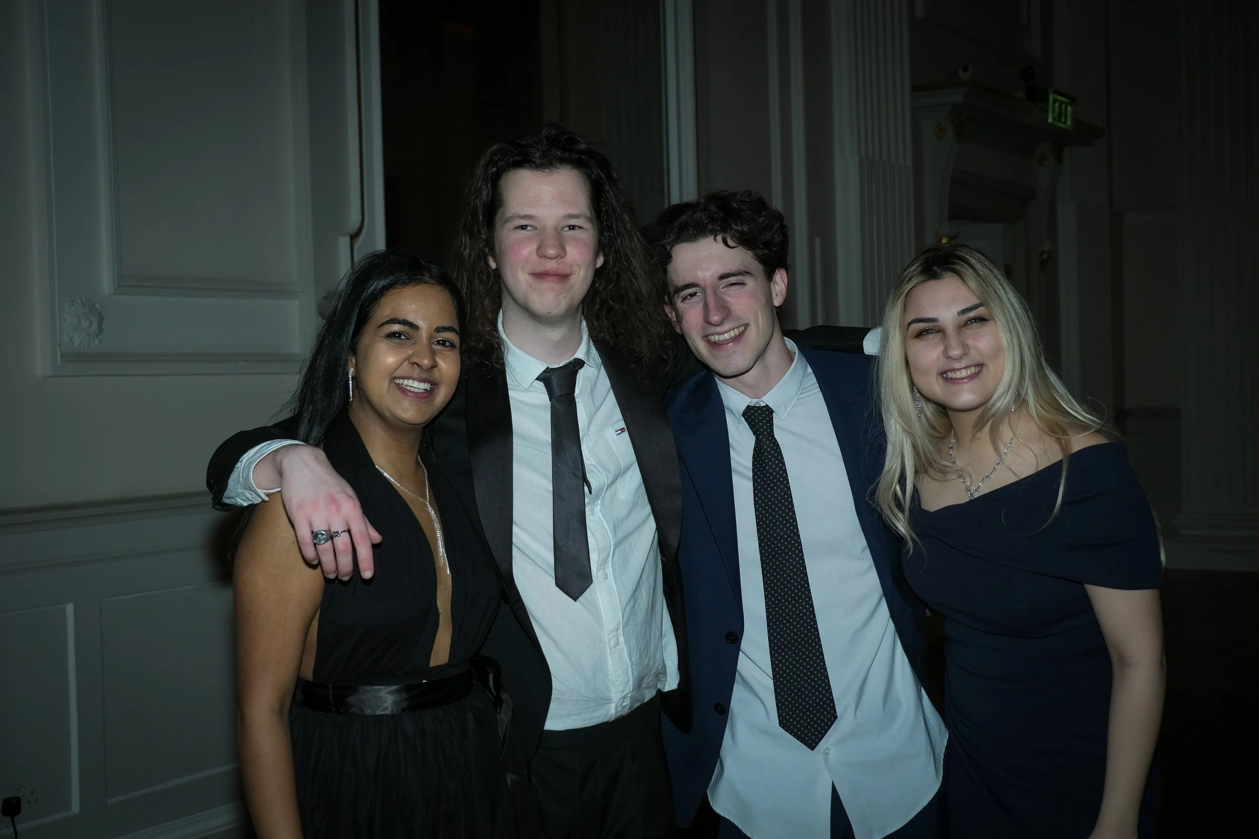 Group of four young adults smiling together at an indoor event, dressed in formal attire.