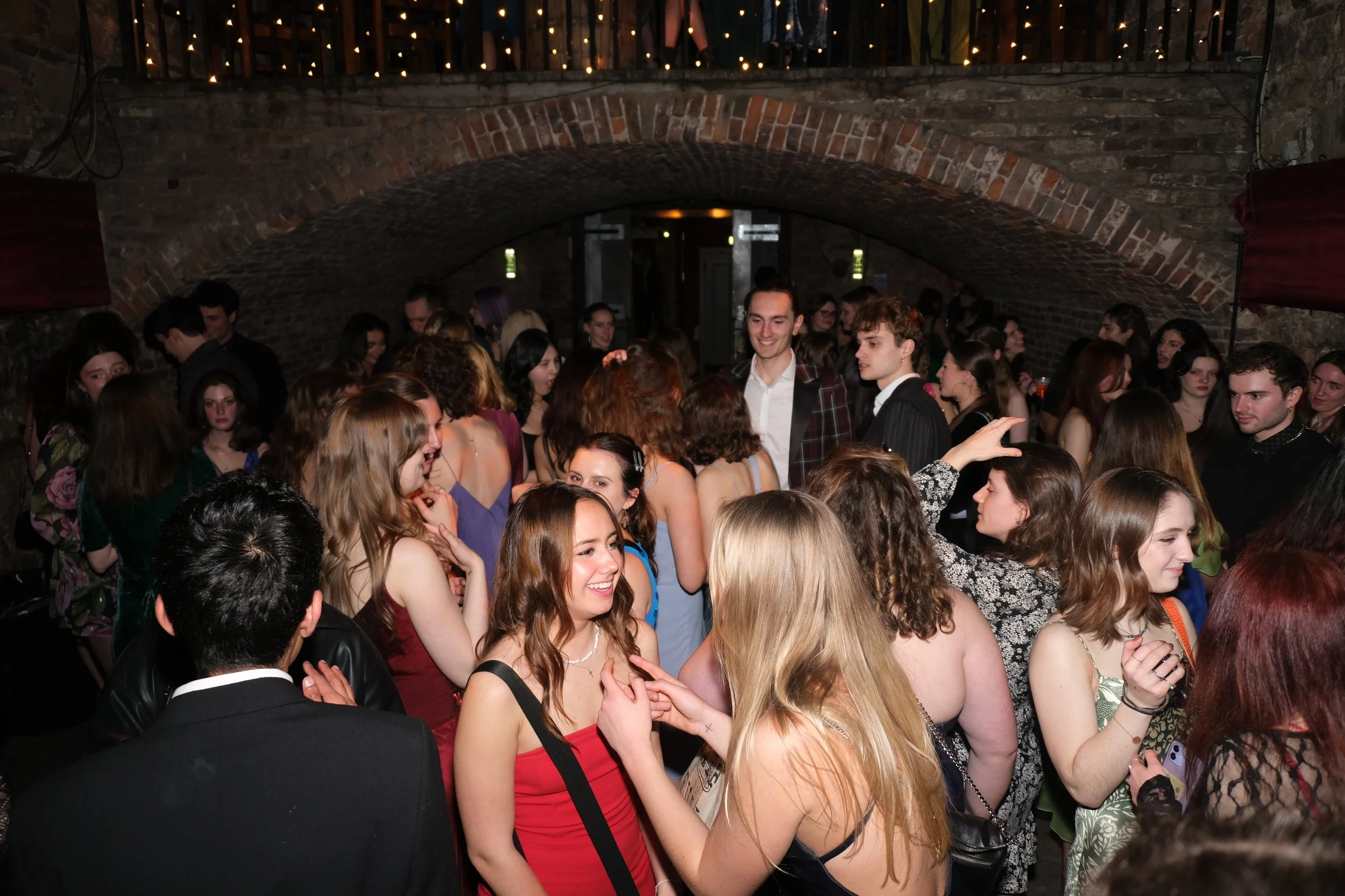 Group of young people at a crowded party or dance in a brick-walled cellar with warm lighting.