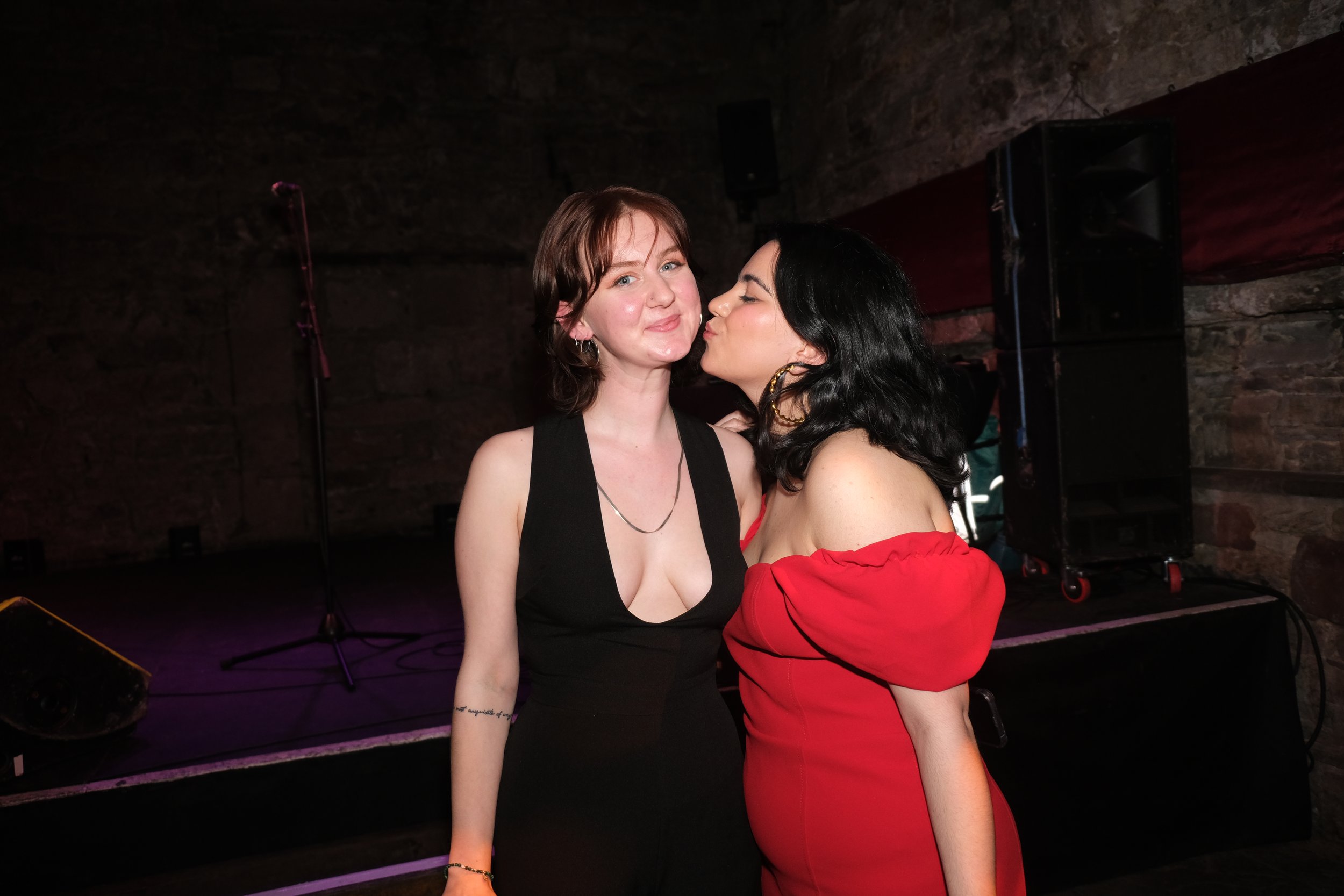 Two women at a party, one in a black sleeveless dress with a deep neckline, the other in a red off-the-shoulder dress, with the second woman leaning in to kiss the first woman on the cheek, against a dark brick wall background with stage equipment.