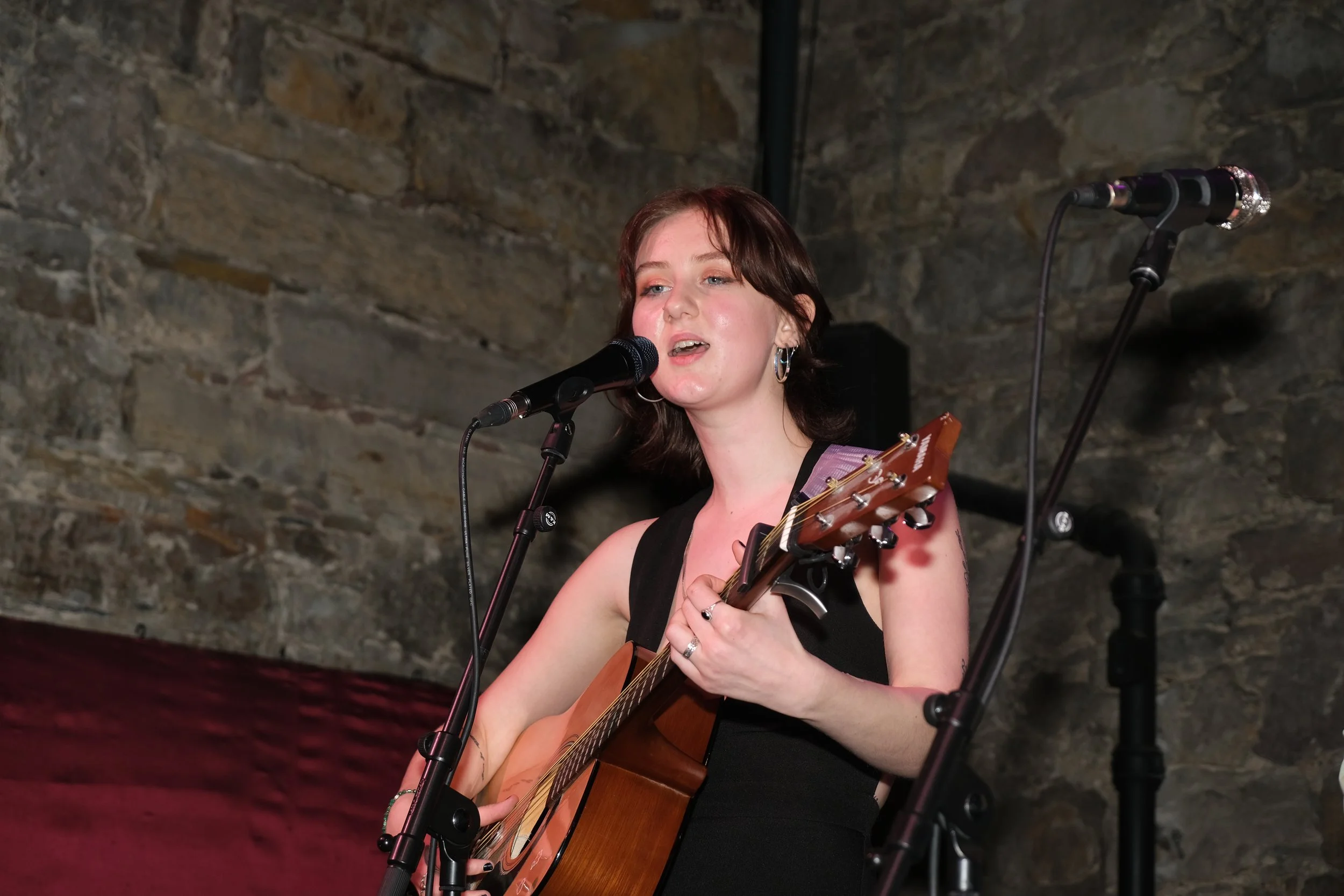 A woman with short brown hair singing into a microphone while playing an acoustic guitar on stage with a stone wall background.