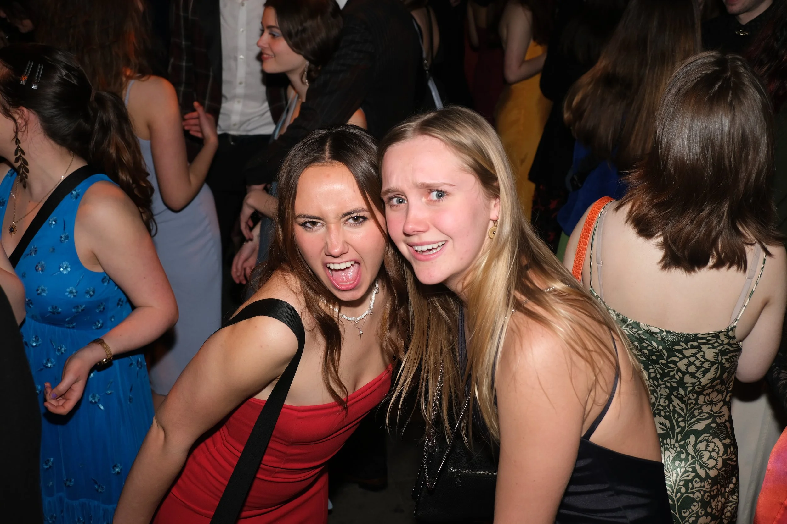 Two young women are making expressive faces at a party or social gathering. Several other people are in the background.