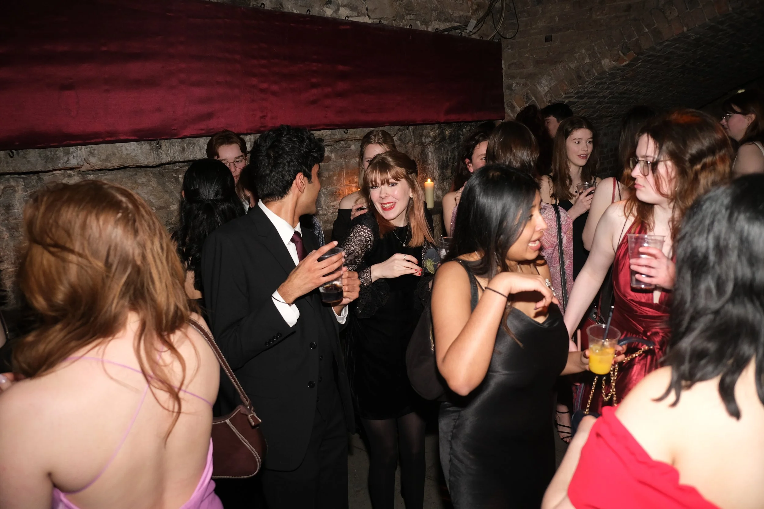 Group of people at a party or social gathering in a dimly lit basement with brick walls, some holding drinks and chatting.