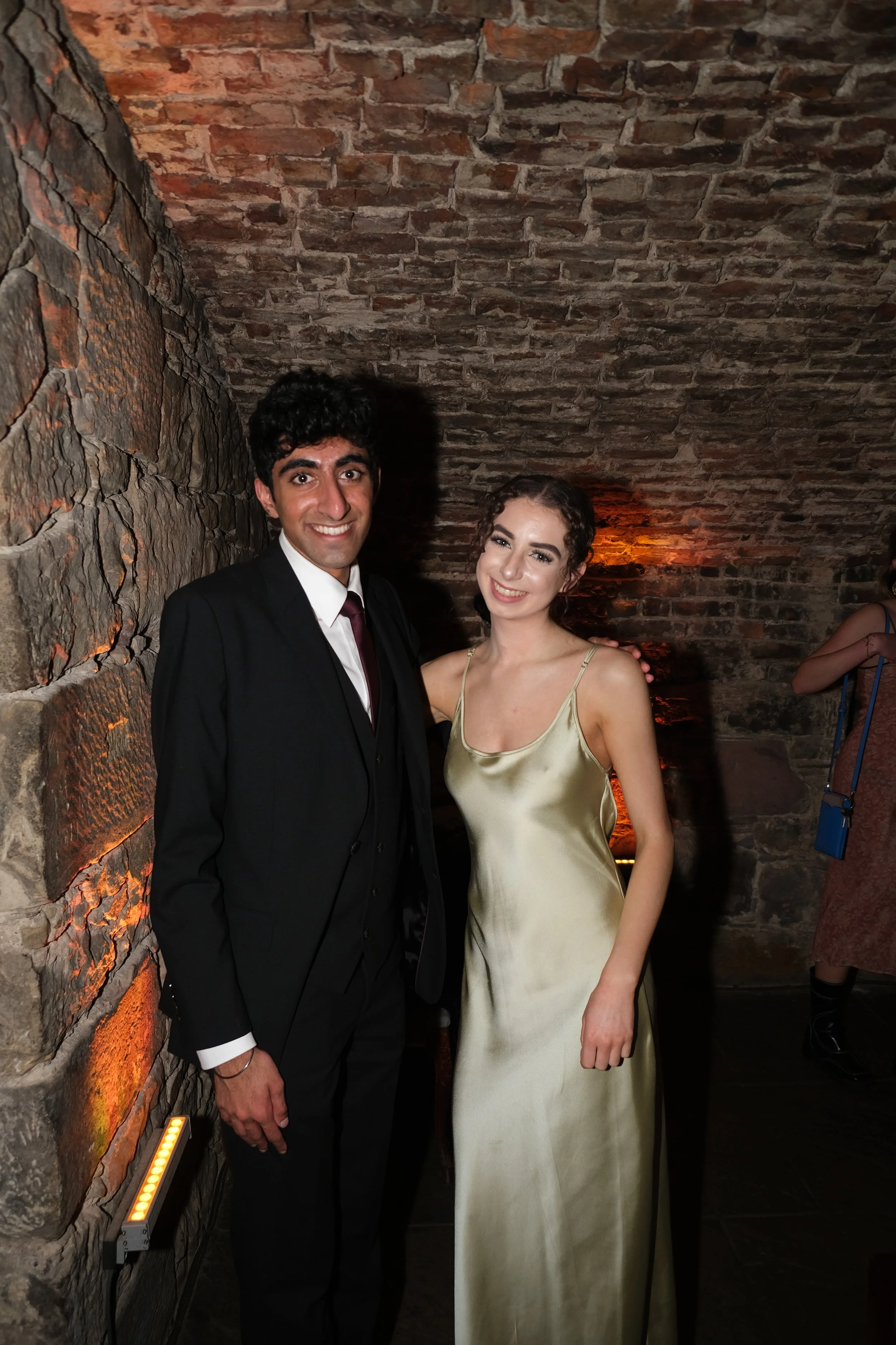 A man in a black suit and a woman in a satin dress pose together in a dimly lit room with exposed brick walls.