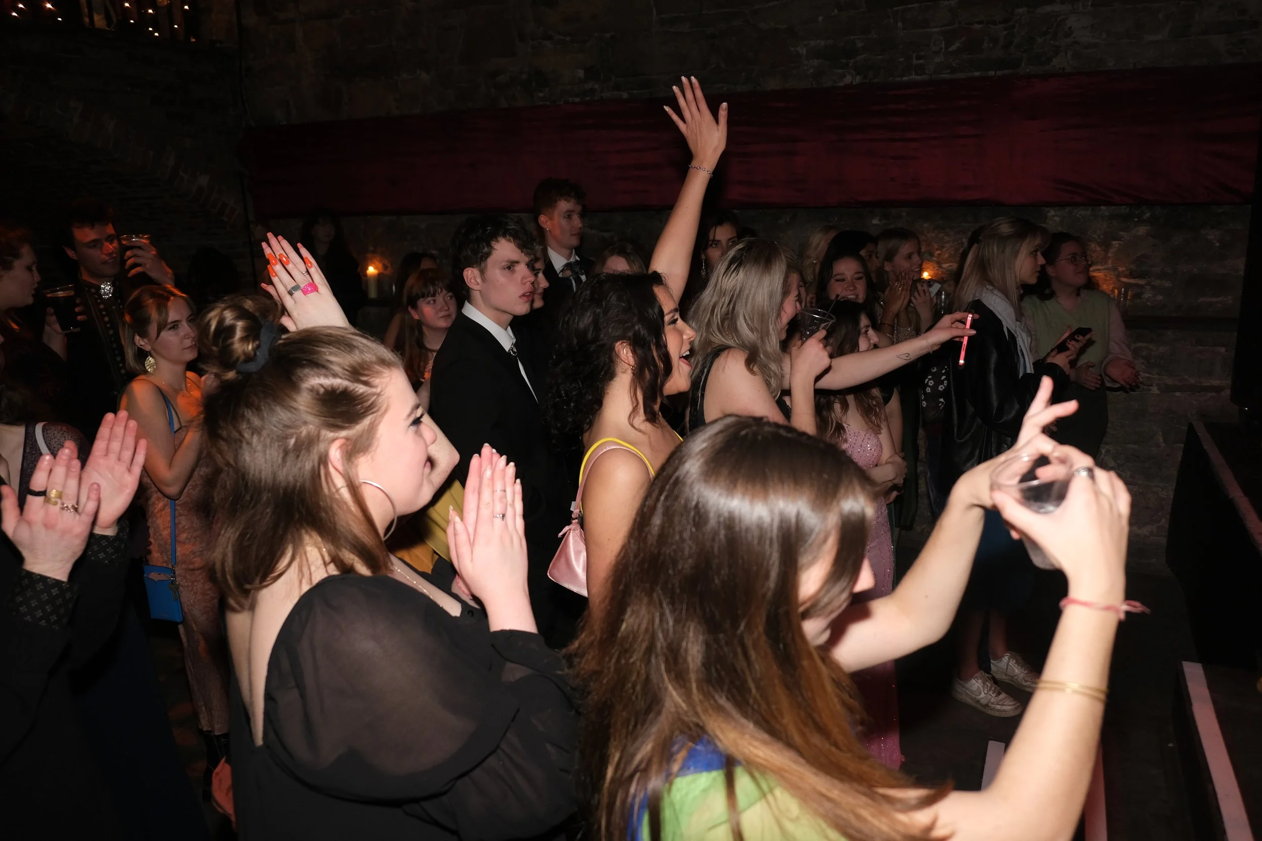 A crowd of young people at a concert or event, some raising their hands and holding drinks, in a dimly lit venue with stone walls.