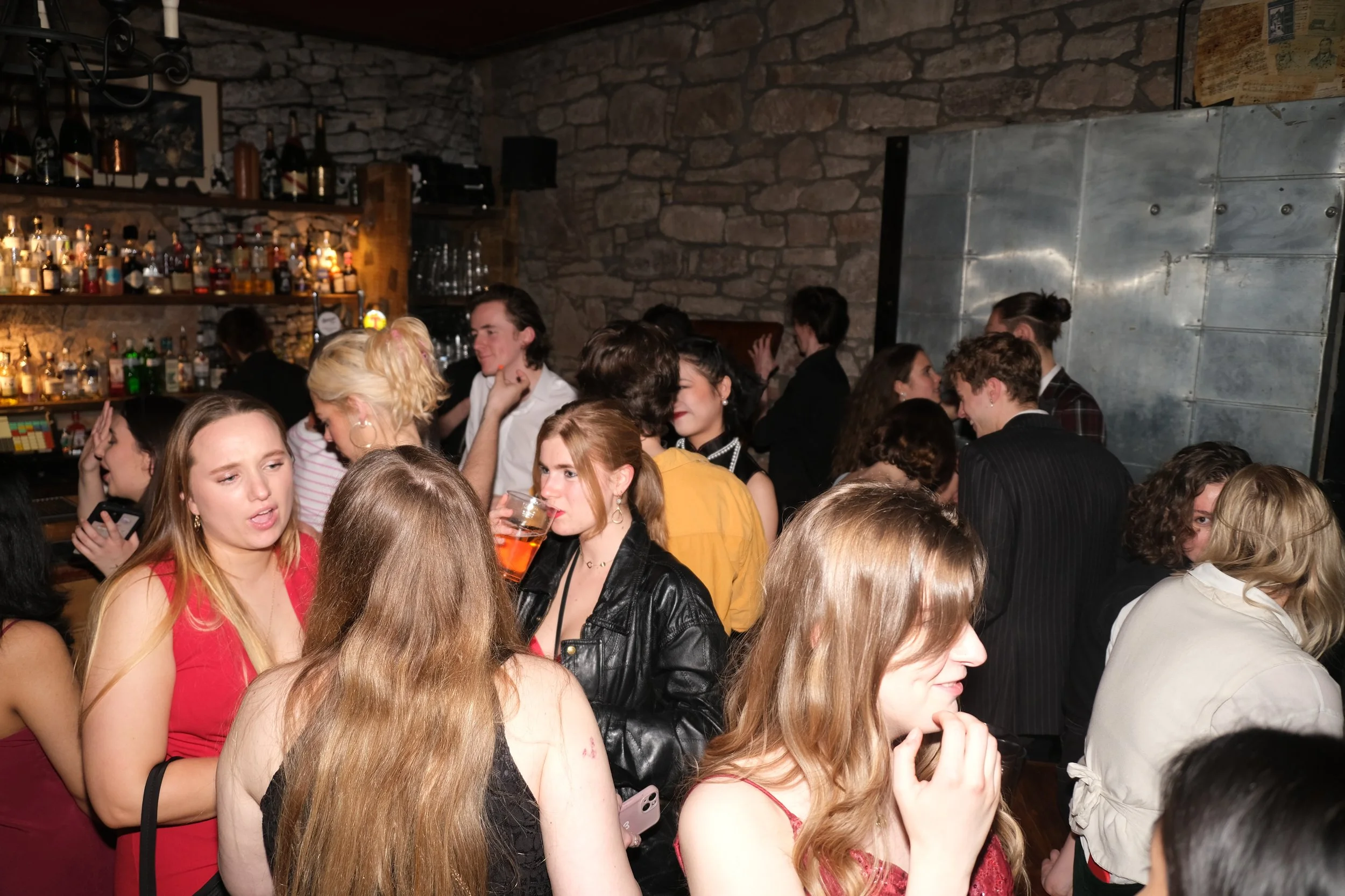 A crowded bar or party scene with people socializing, chatting, and holding drinks in a dimly lit setting with stone walls and liquor bottles on shelves.