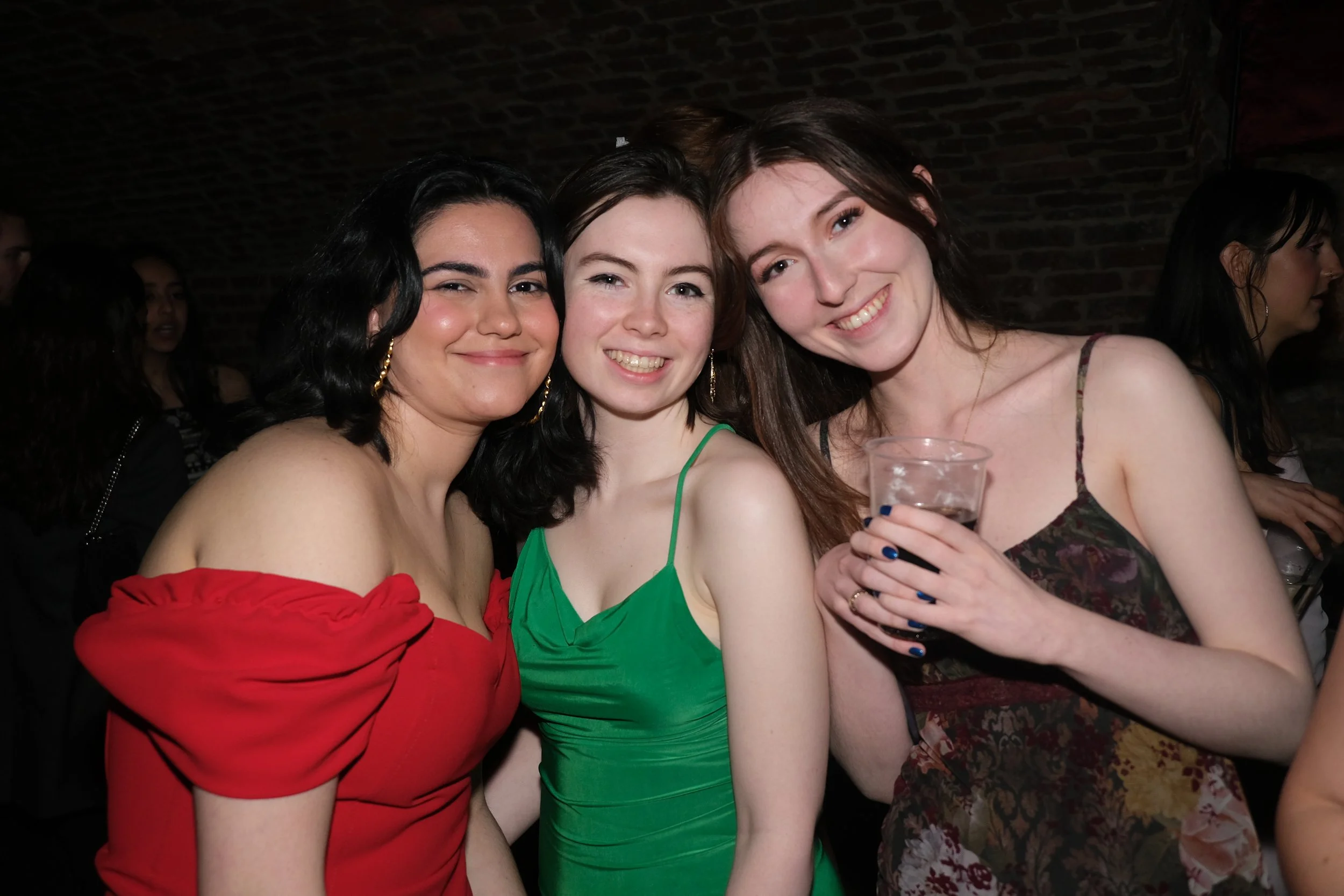Three women smiling and posing for a photo at a party or social event, with one woman holding a drink
