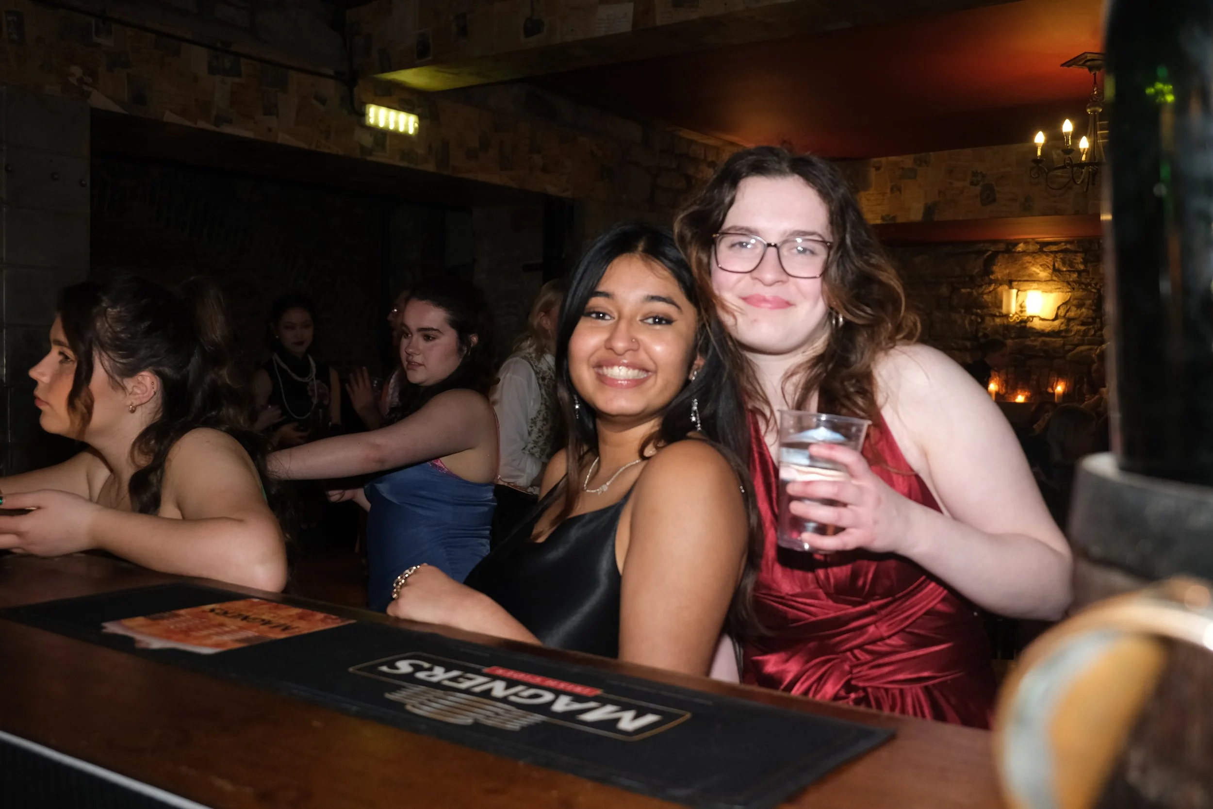 Two women smiling at a bar, one holding a glass of water, with other women in the background, in a dimly lit restaurant or bar setting.