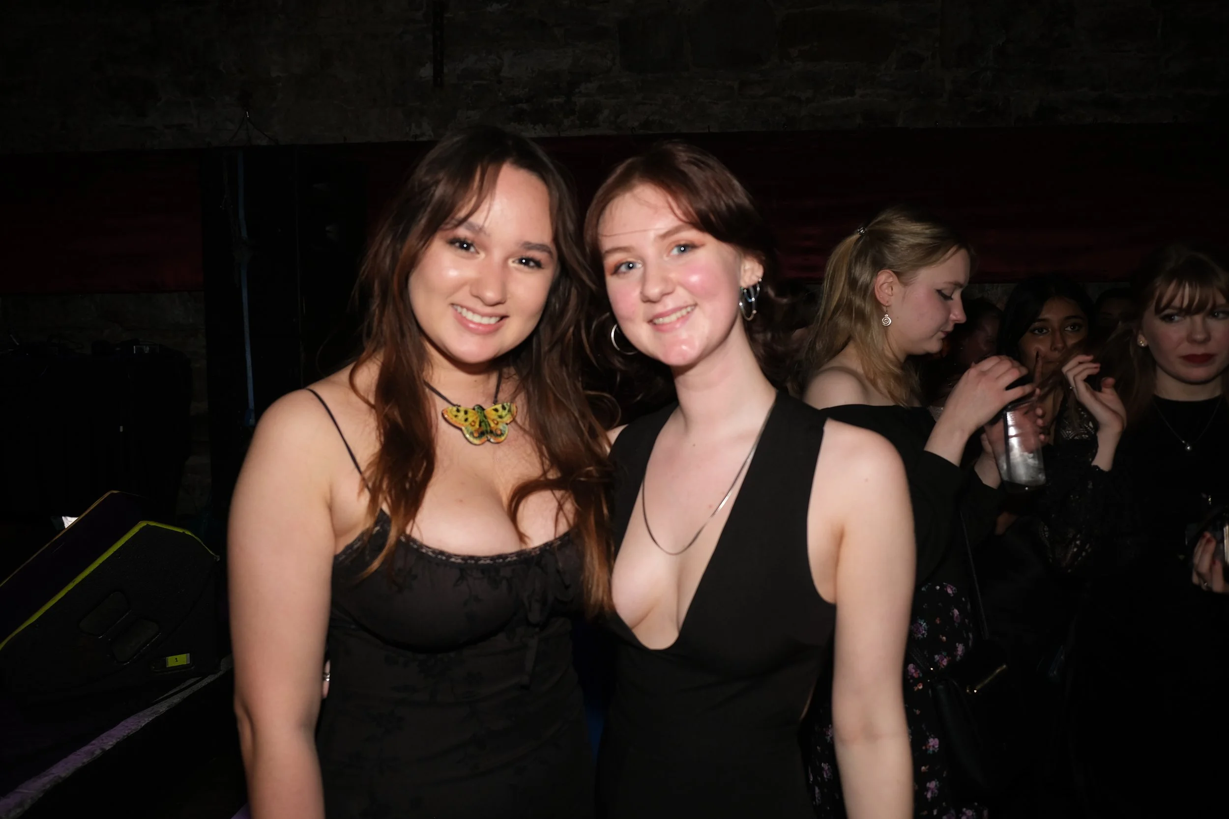 Two young women in black dresses smiling and posing together at a social event, with a group of women in the background holding drinks.