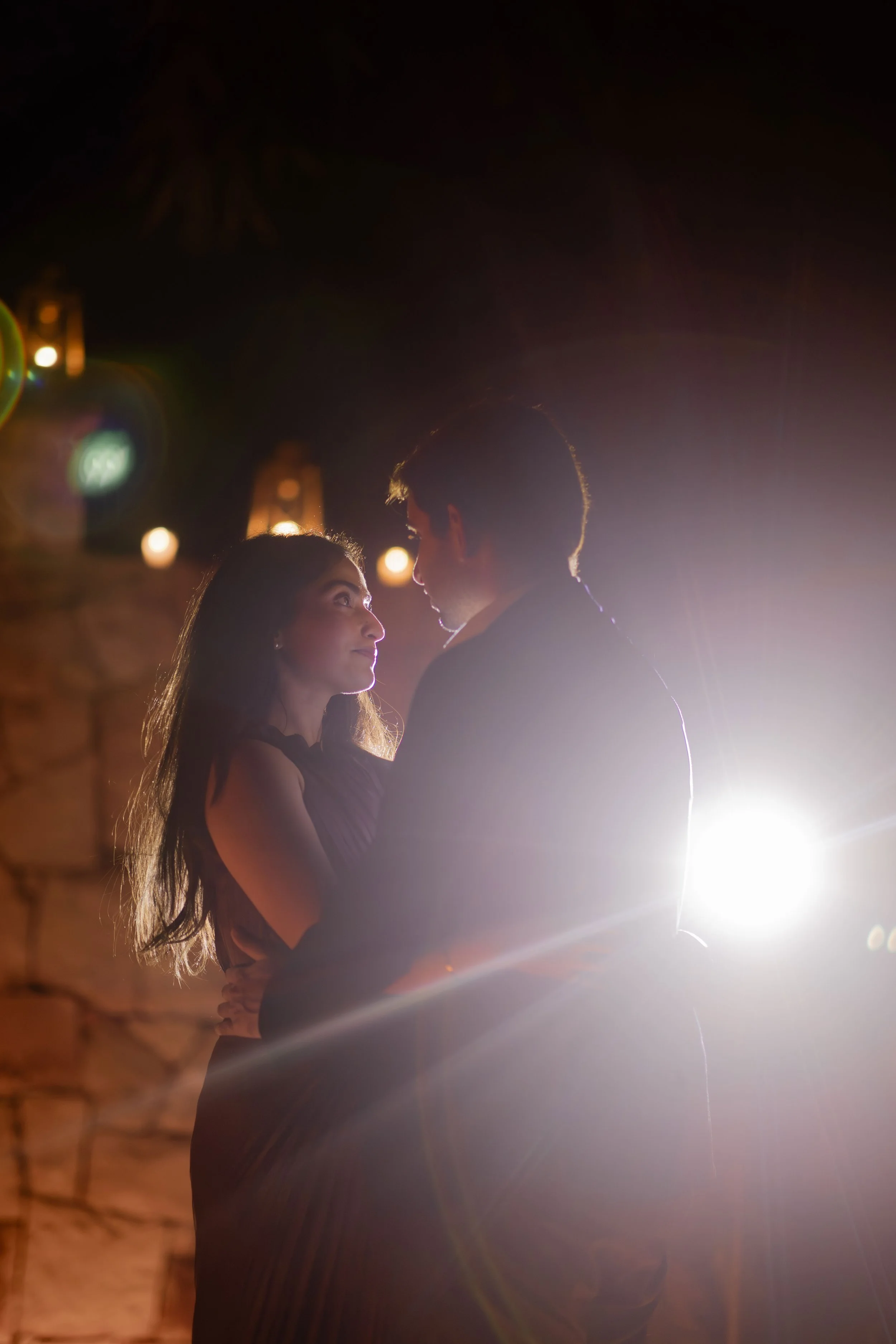 A couple dancing closely in a dimly lit setting, with warm lighting and a bright backlight illuminating their profiles.