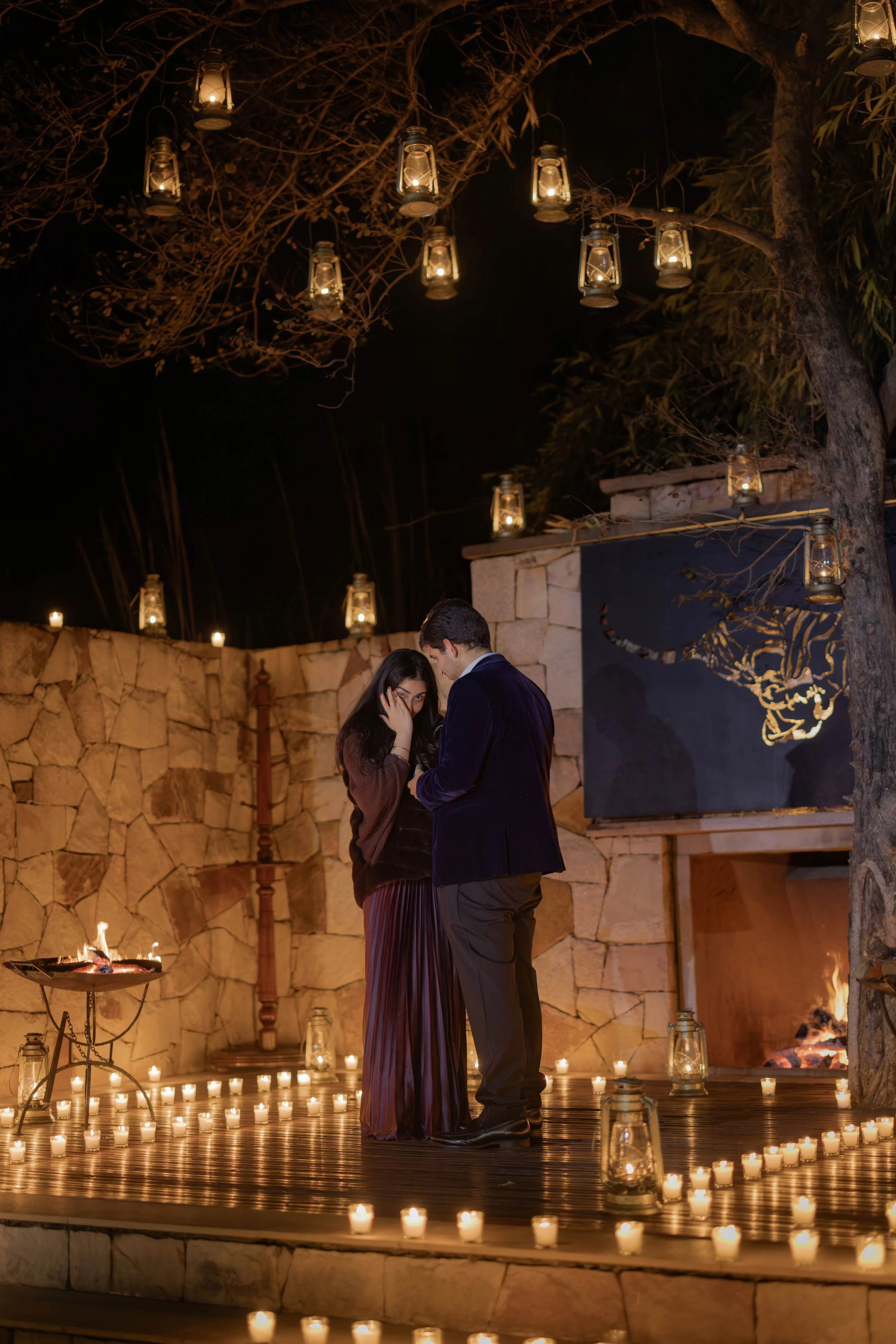A couple standing close together surrounded by candles and lanterns in an outdoor setting at night.