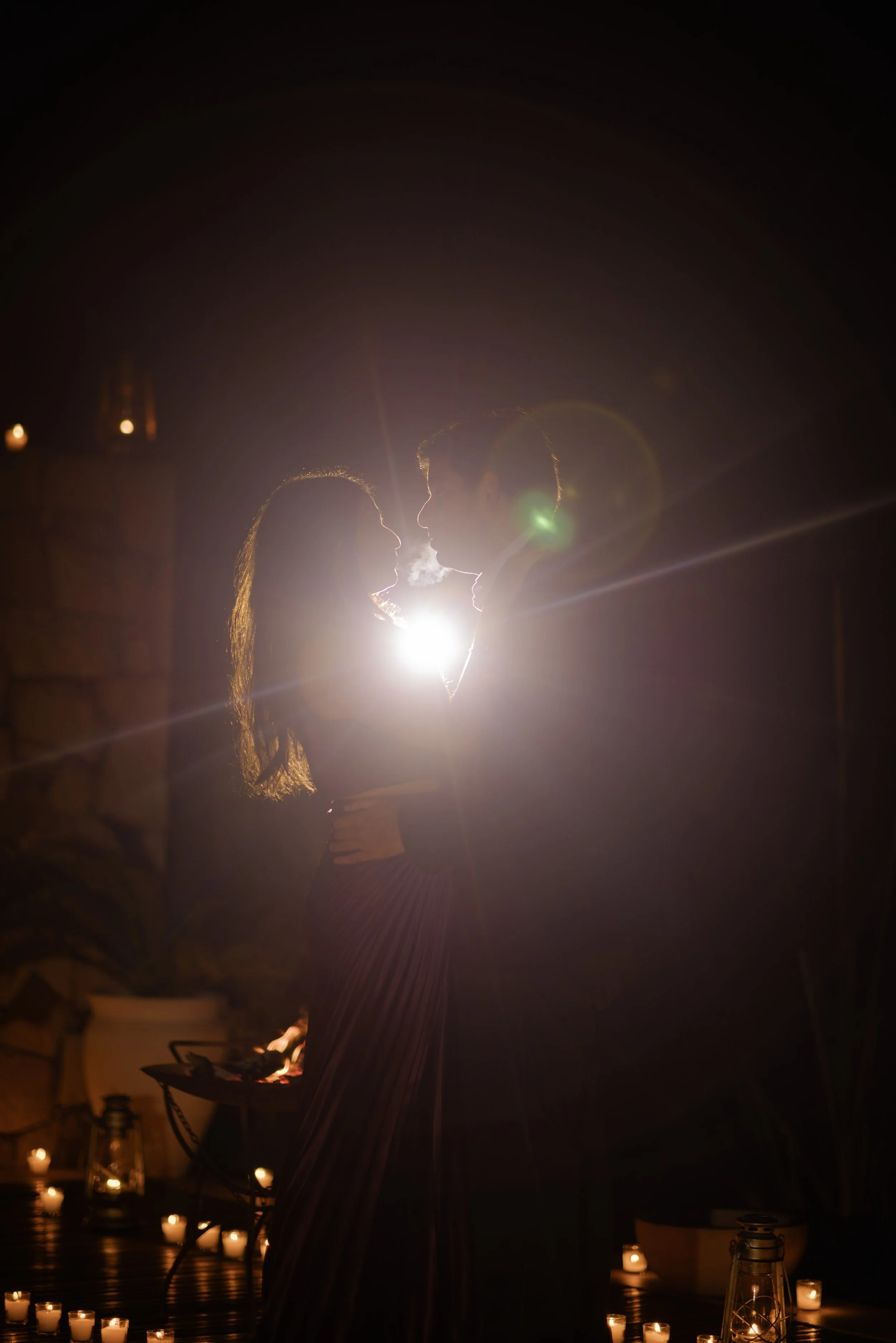 A silhouette of a couple embracing in a dimly lit room with candles, with a bright light behind them creating a halo effect.