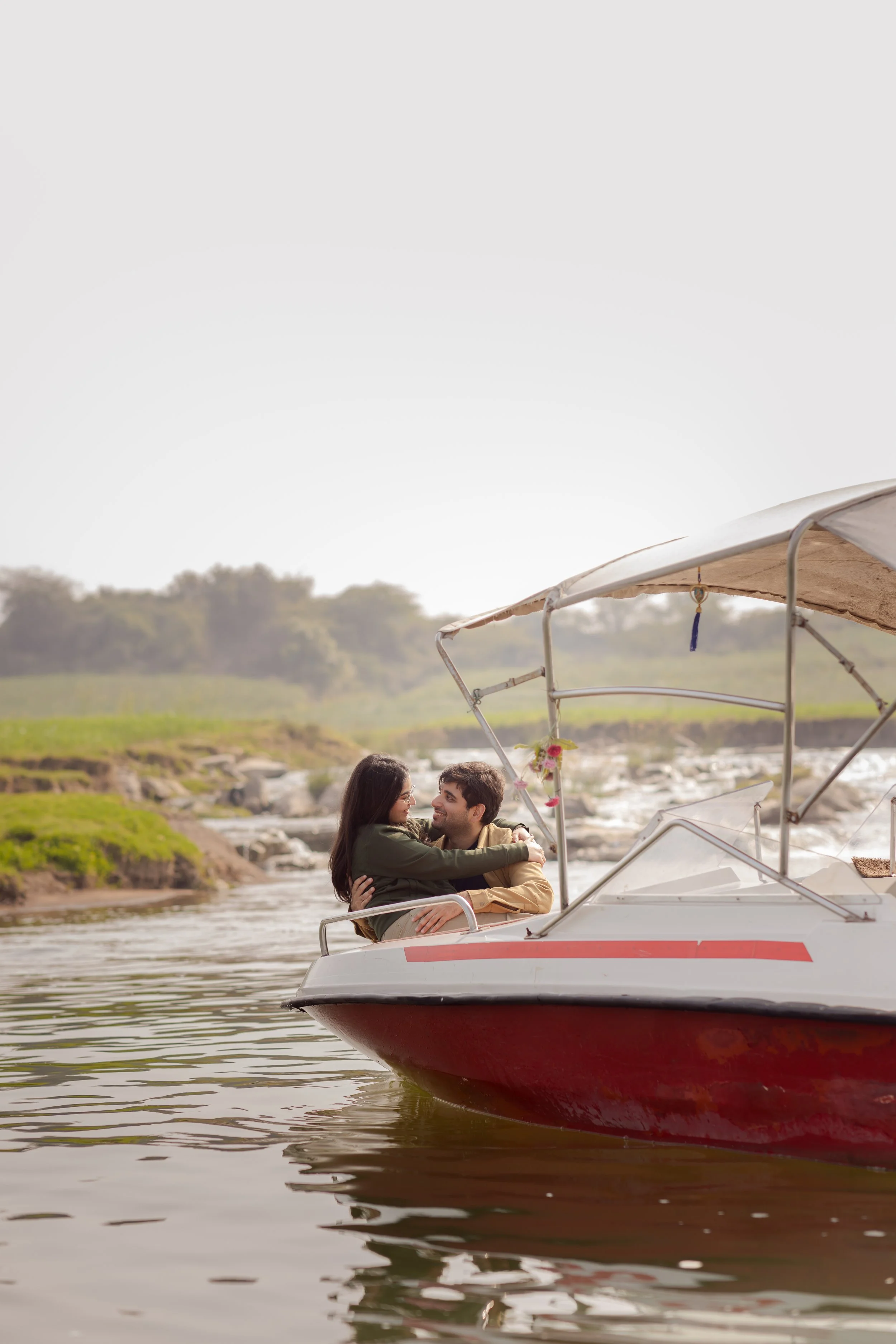 A couple embraces on a boat floating on a calm river, with green wooded hills in the background.