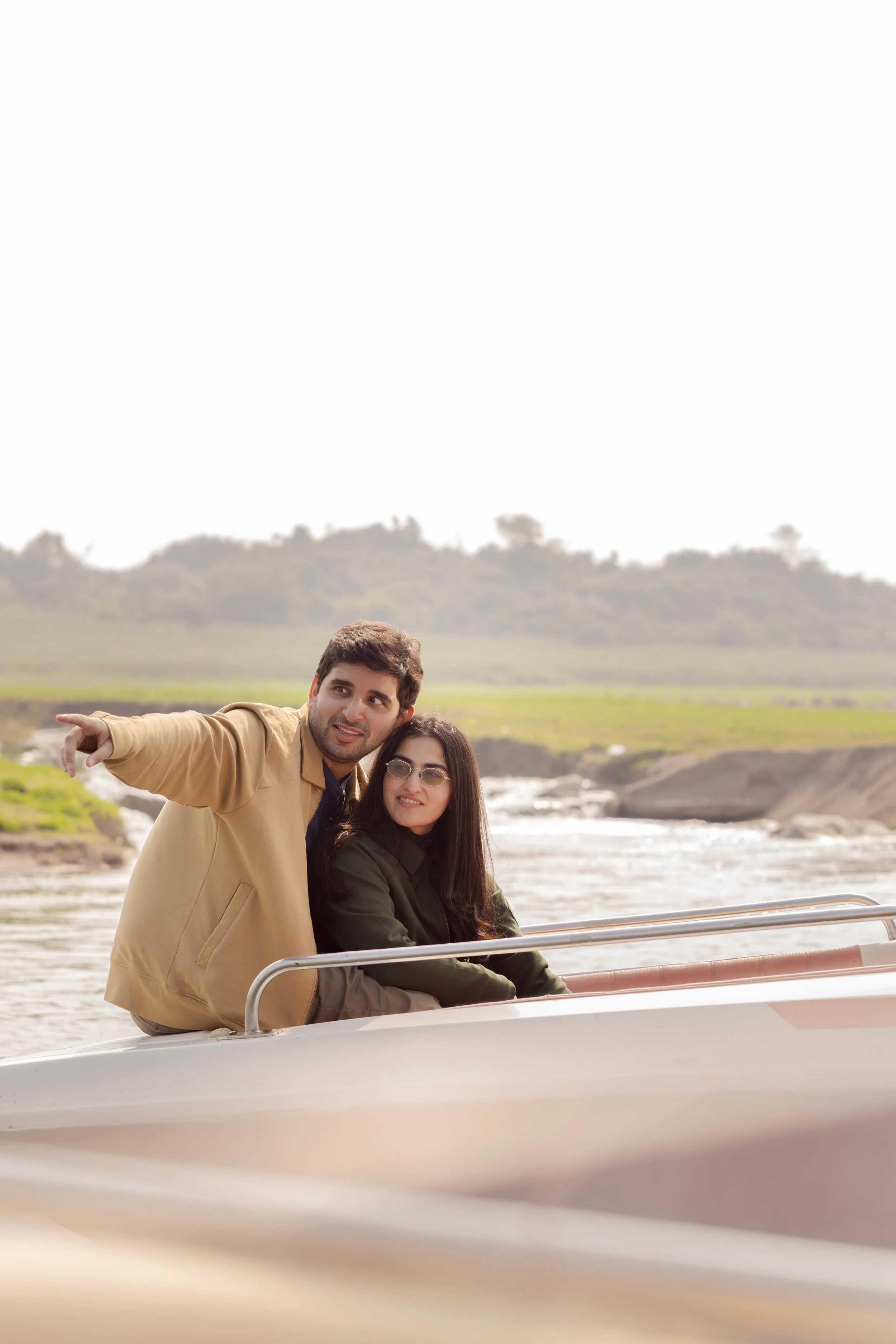 A man and woman on a boat, with the man pointing at something in the distance, in a scenic outdoor setting with water and greenery.