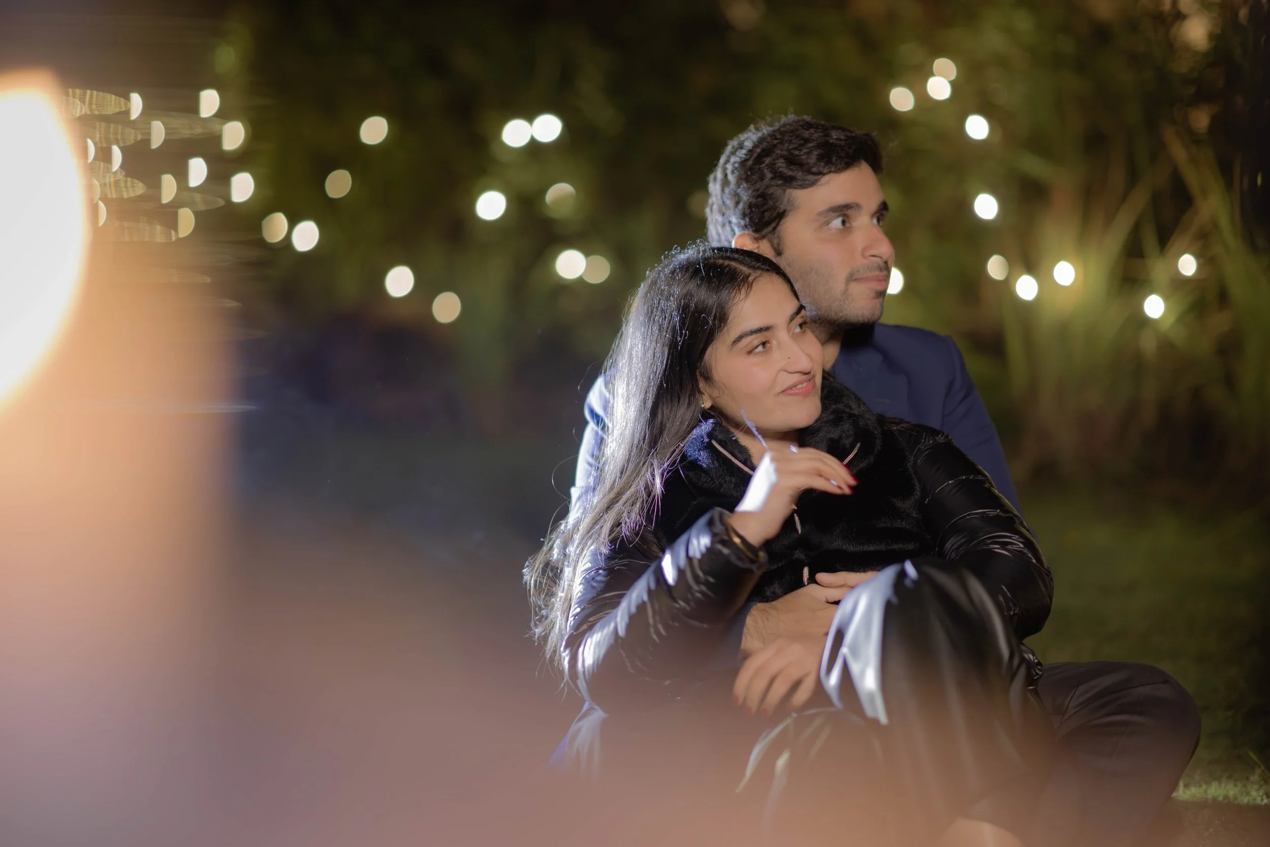 A young couple sitting outdoors at night, surrounded by blurred string lights and greenery, with the man embracing the woman from behind.