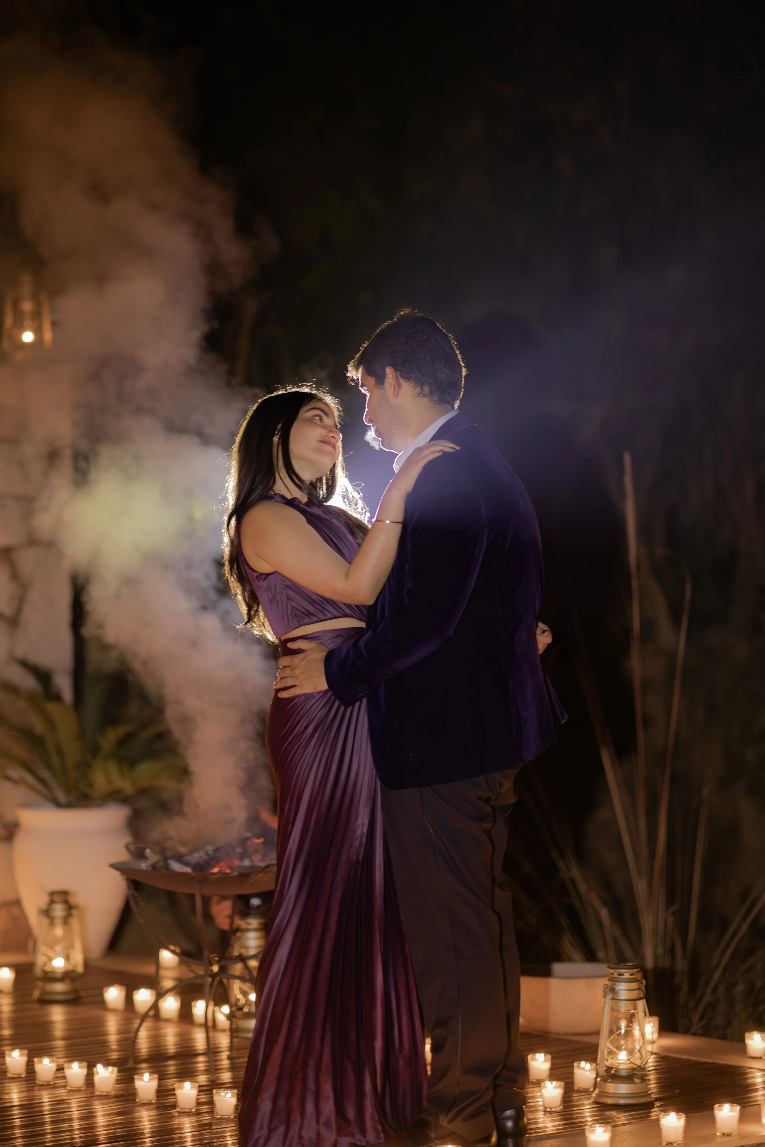A couple dancing close together outdoors at night with candlelights and lanterns around, under a misty, dark background.