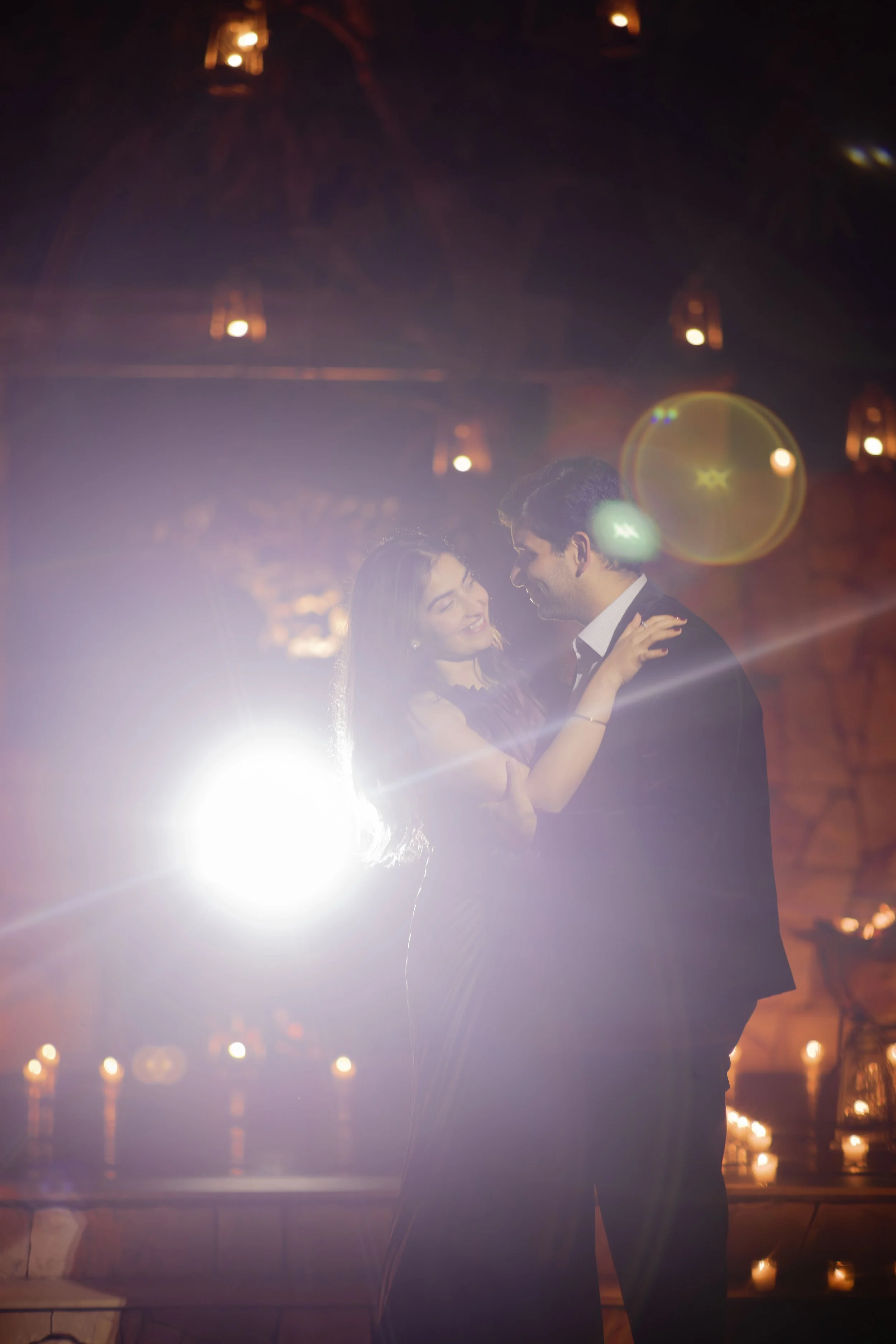 A couple dancing closely at an indoor event, illuminated by a bright backlight and warm ambient lighting.