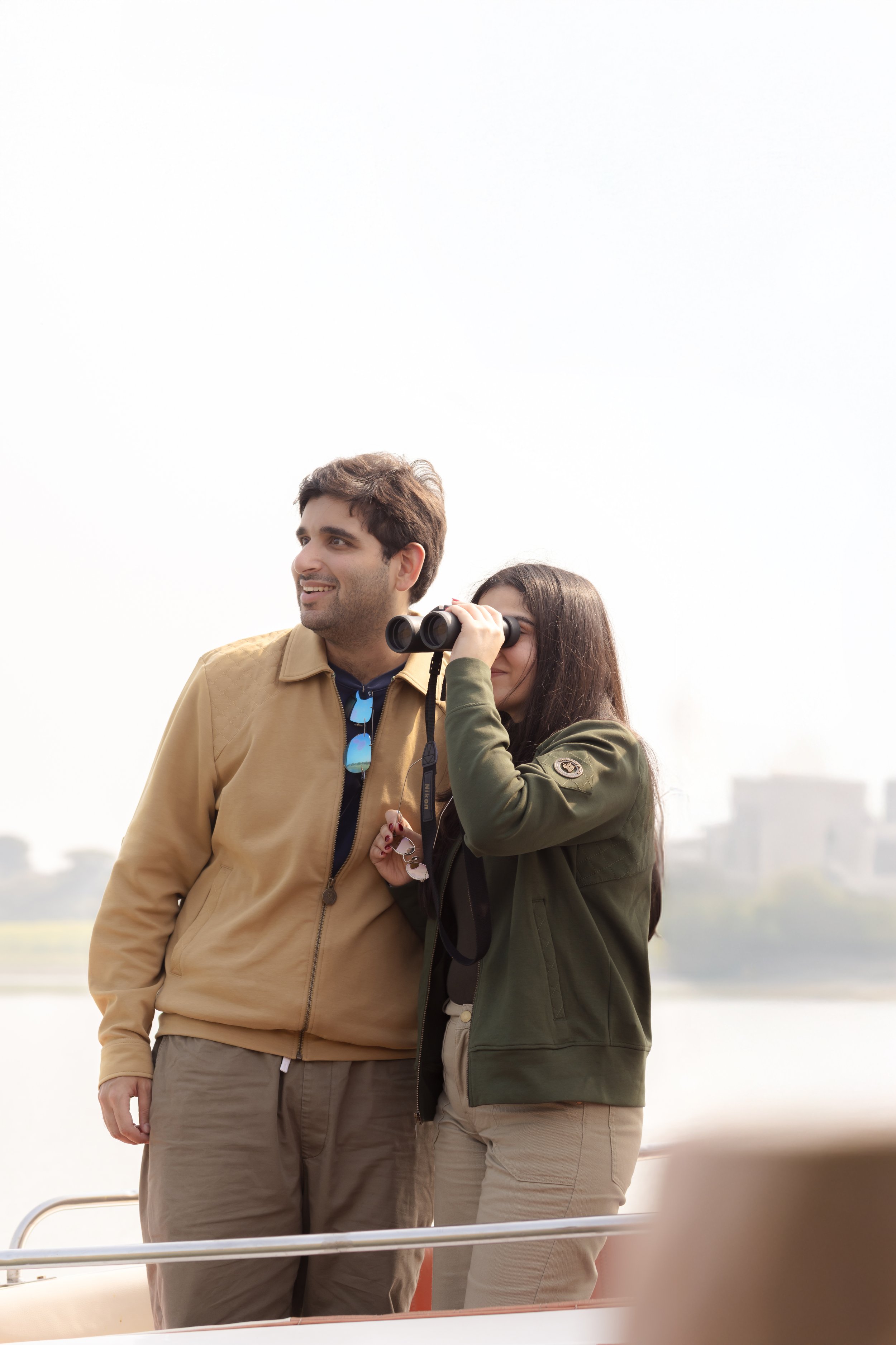 A man and woman standing outdoors, with the woman looking through binoculars, both smiling and enjoying the view.