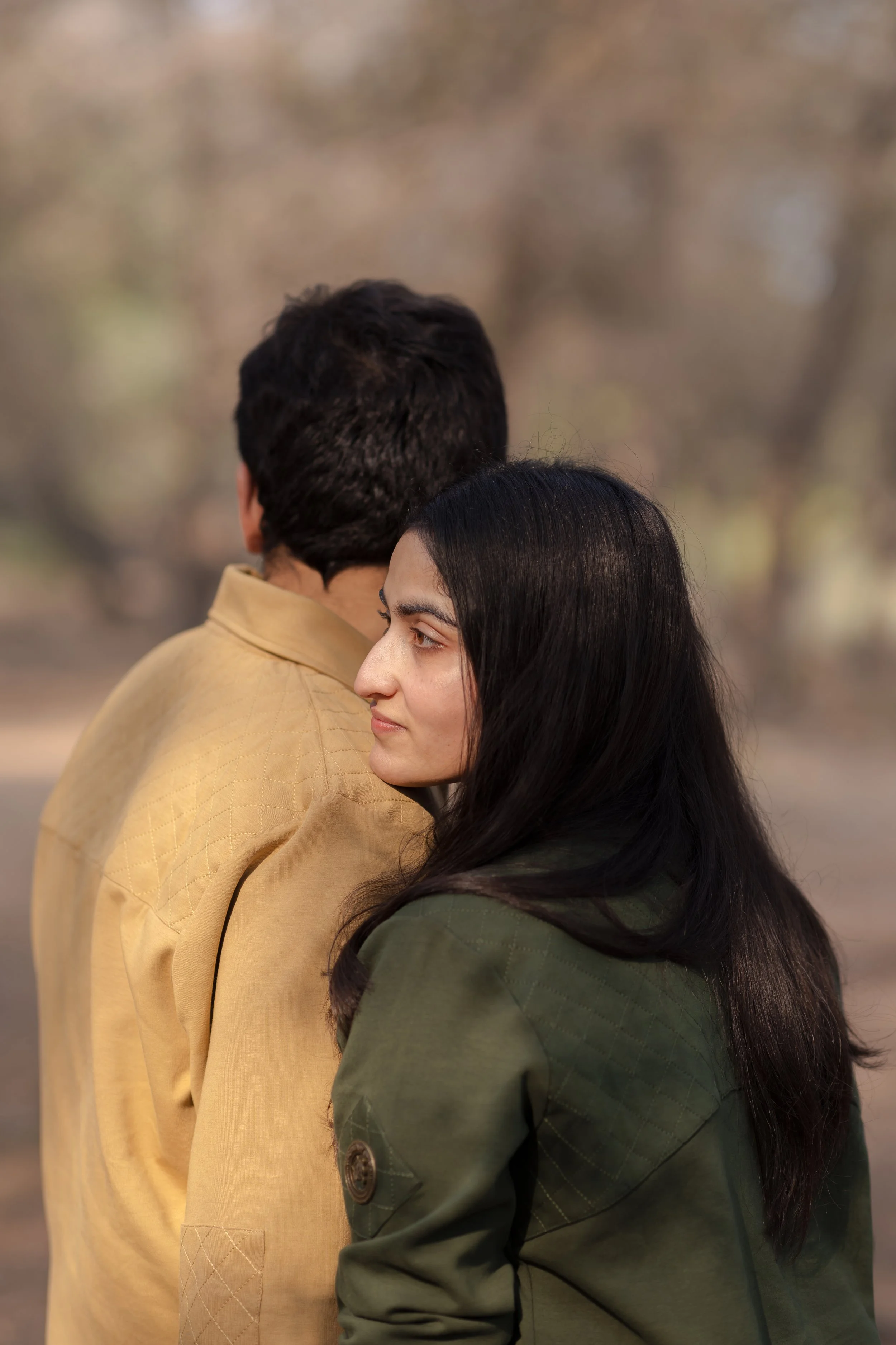 A young woman with long dark hair and a man with short dark hair outdoors, with trees in the background. The woman is leaning on the man's shoulder, both facing sideways.