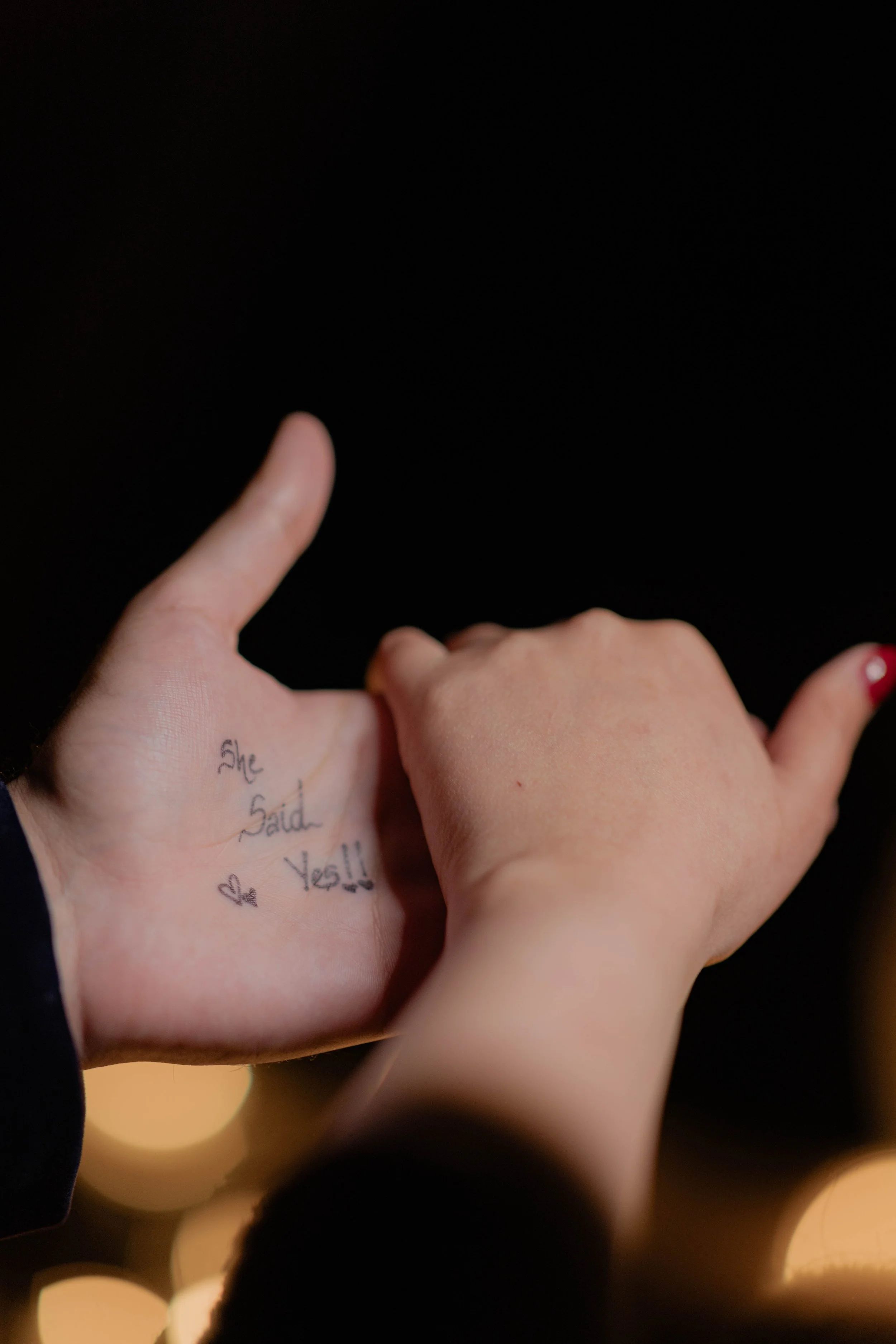Close-up of a hand with a tattoo that reads 'She Said Yes!!' and a small heart. The hand is making a shaka sign against a dark background with bokeh lights.