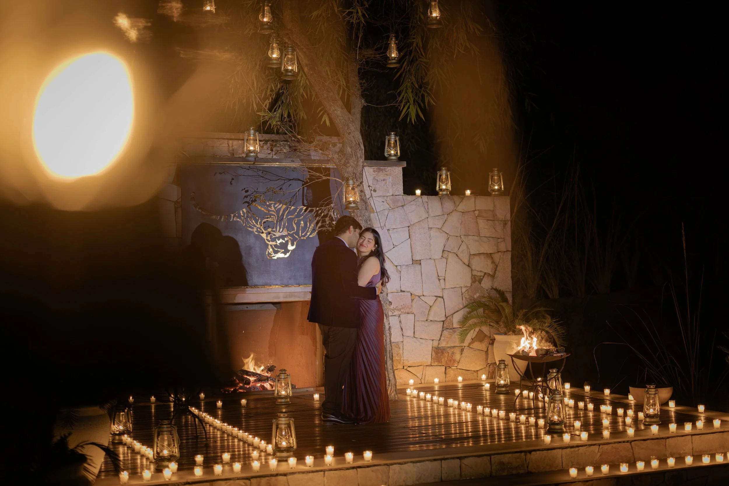 A couple dancing during a romantic outdoor event at night, illuminated by candles and lanterns with a fireplace and stone wall in the background.