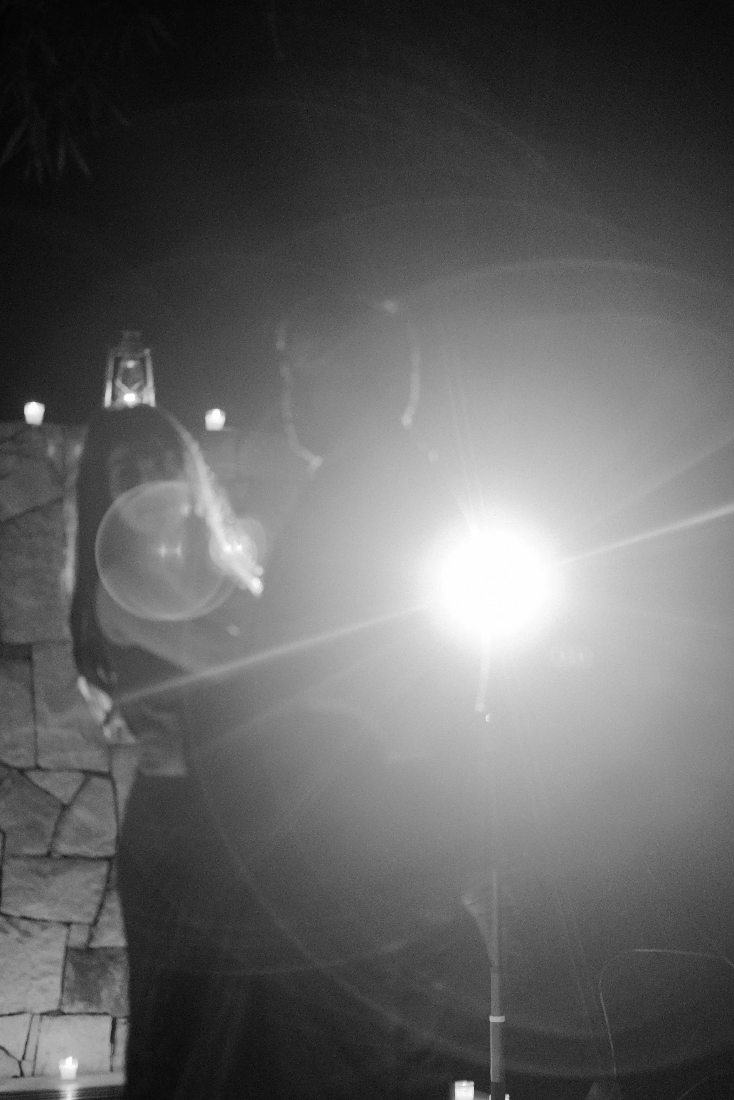 Silhouettes of a man and woman standing outdoors at night illuminated by a bright light, with candles and a stone wall in the background.