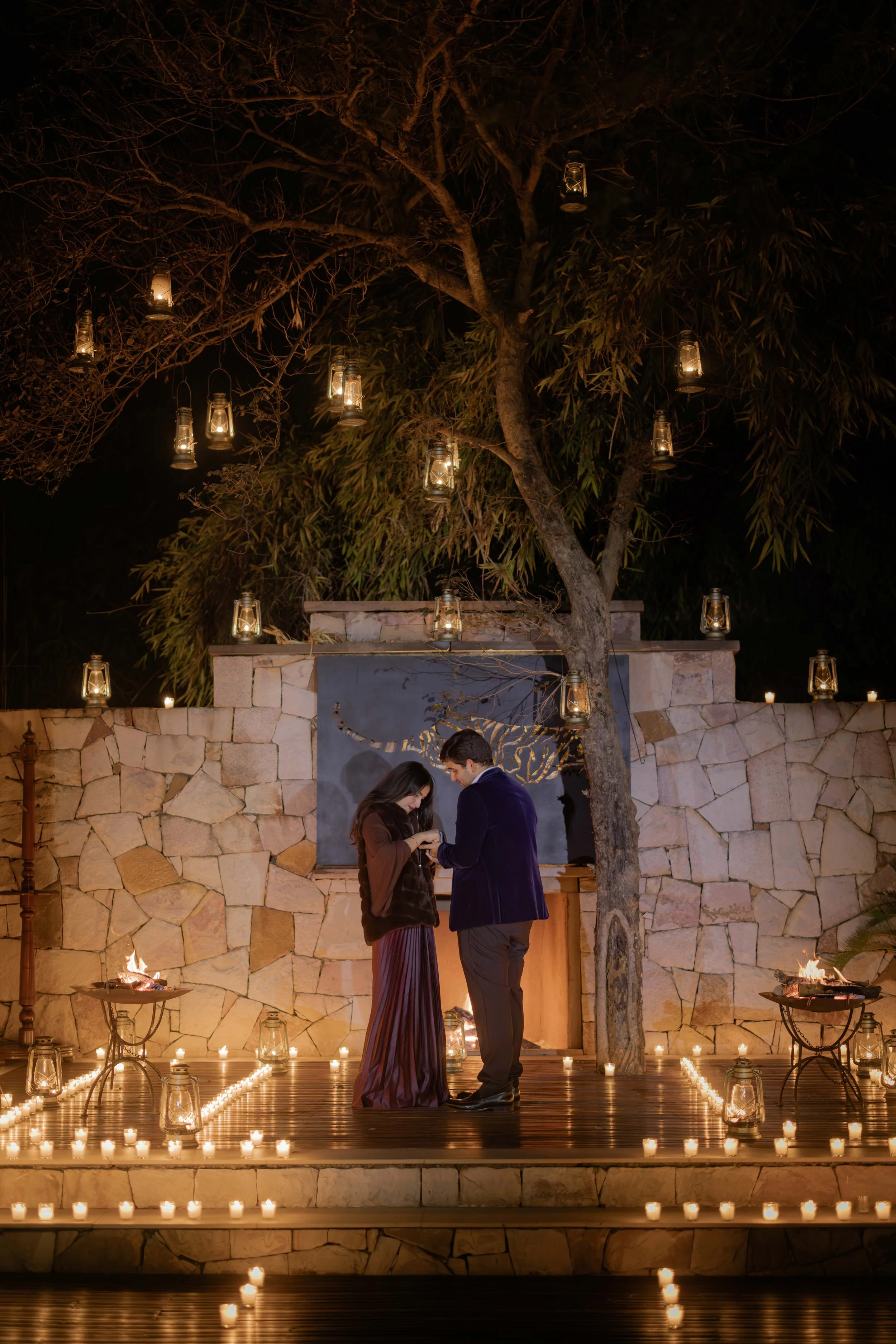 A couple is standing close together on a wooden deck surrounded by candlelight at night. The setting includes a stone wall, a large tree with hanging lanterns, and small fires in torches, creating a romantic ambiance.