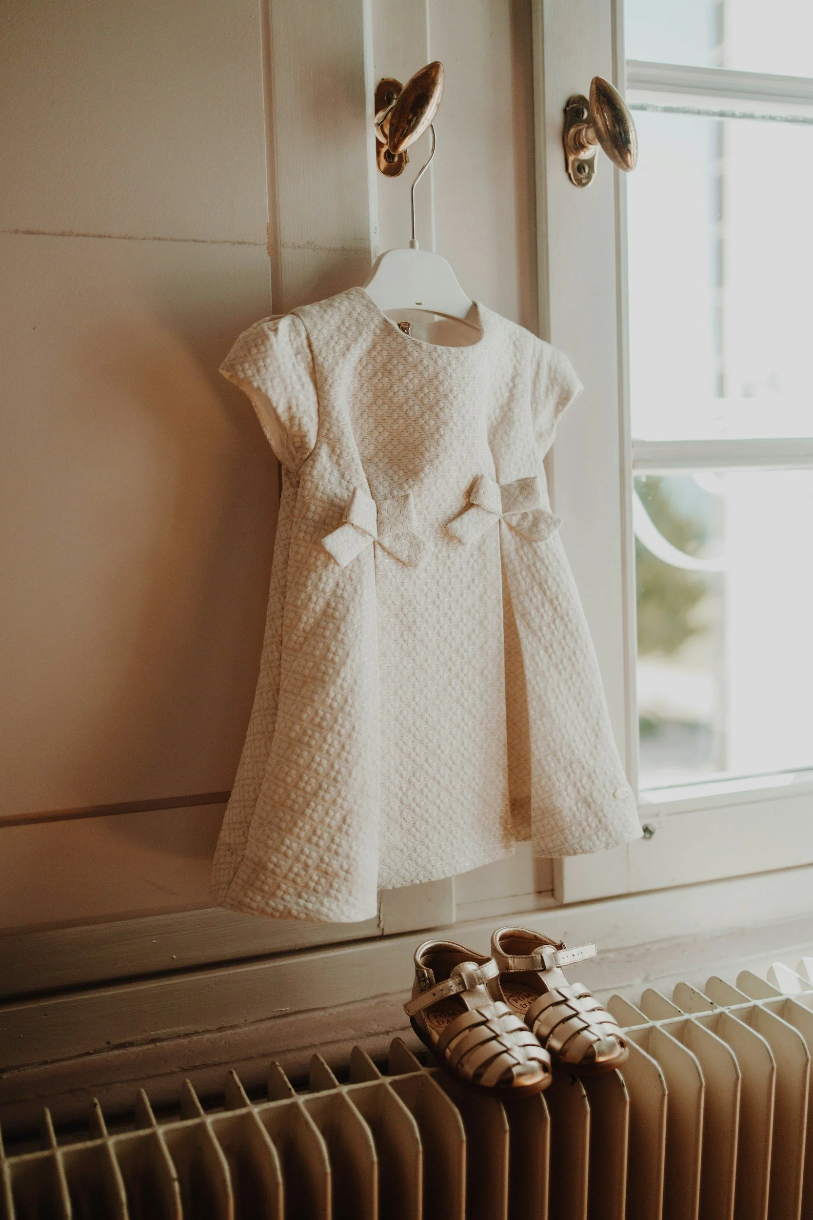 White textured baby dress hanging beside a sunlit window, with small brown sandals on a radiator below
