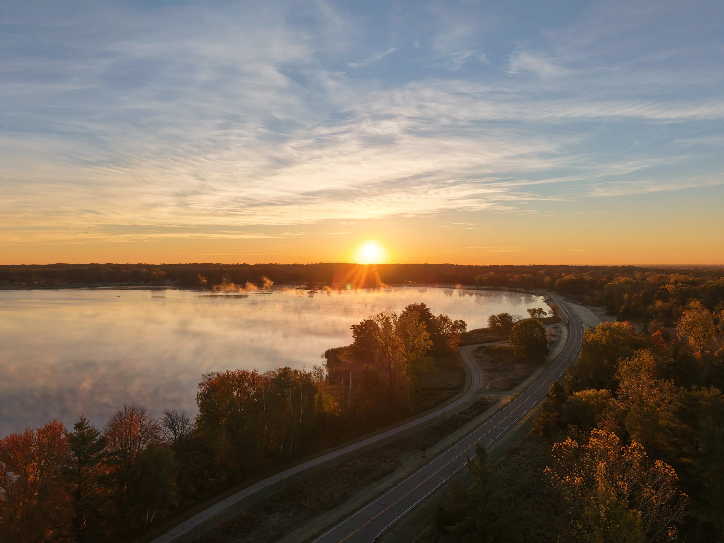 Aerial view of a sunset over a calm lake, winding road, and autumn forest