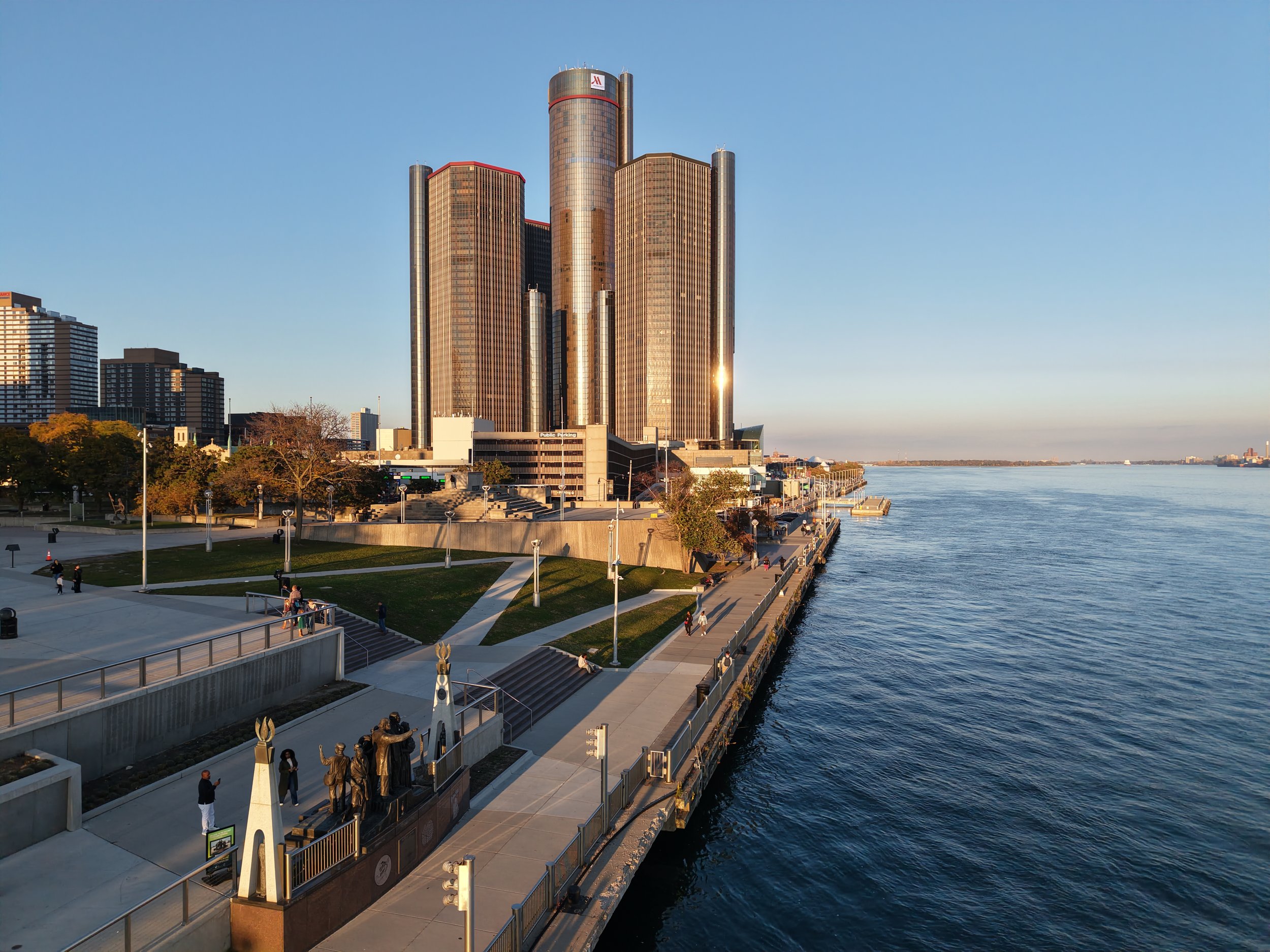 Detroit River waterfront with the Renaissance Center skyscrapers, riverwalk, and public art under a clear blue sky