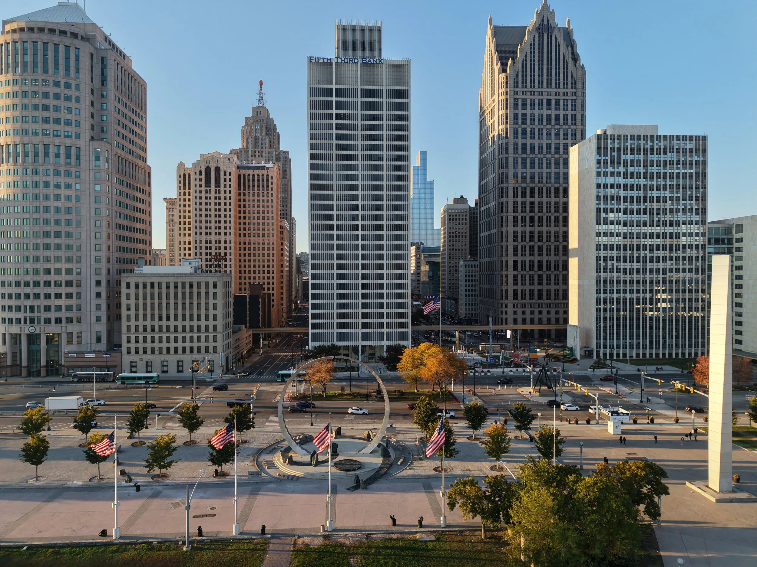 Detroit skyline viewed from Hart Plaza, featuring the Fifth Third Bank building, Ally Detroit Center, traffic, and American flags.