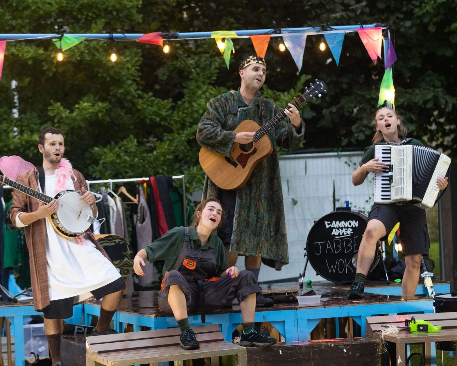 Outdoor theatrical band performing with guitar, banjo, accordion, and drums under colorful bunting and string lights