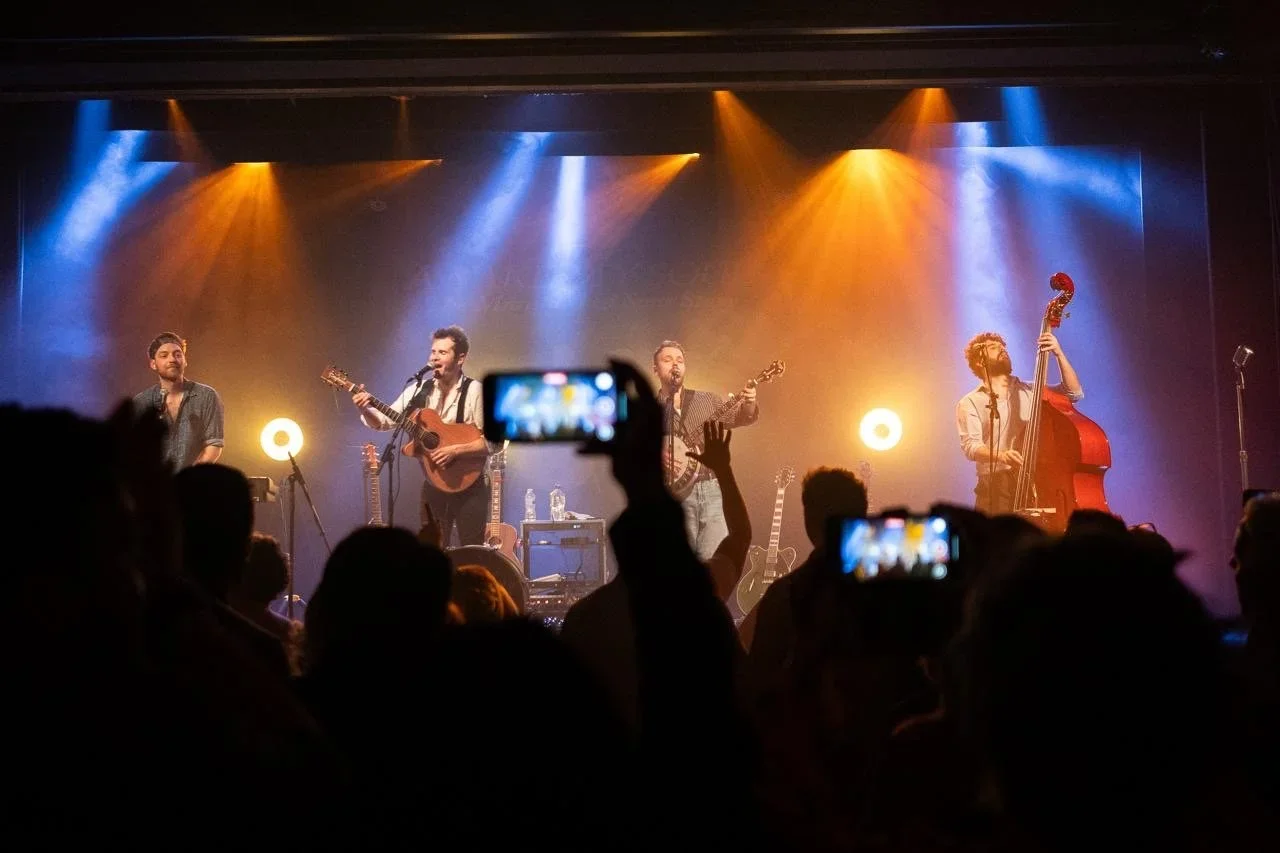 The Mumford & Sons Story on stage under blue and orange lights as a crowd dances and records with smartphones